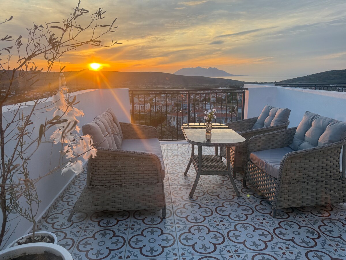 An outdoor terrace is shown with a cozy seating arrangement, featuring two cushioned chairs and a small table. The horizon displays a stunning sunset over distant mountains and the sea, with a delicate plant adding a touch of nature to the scene.