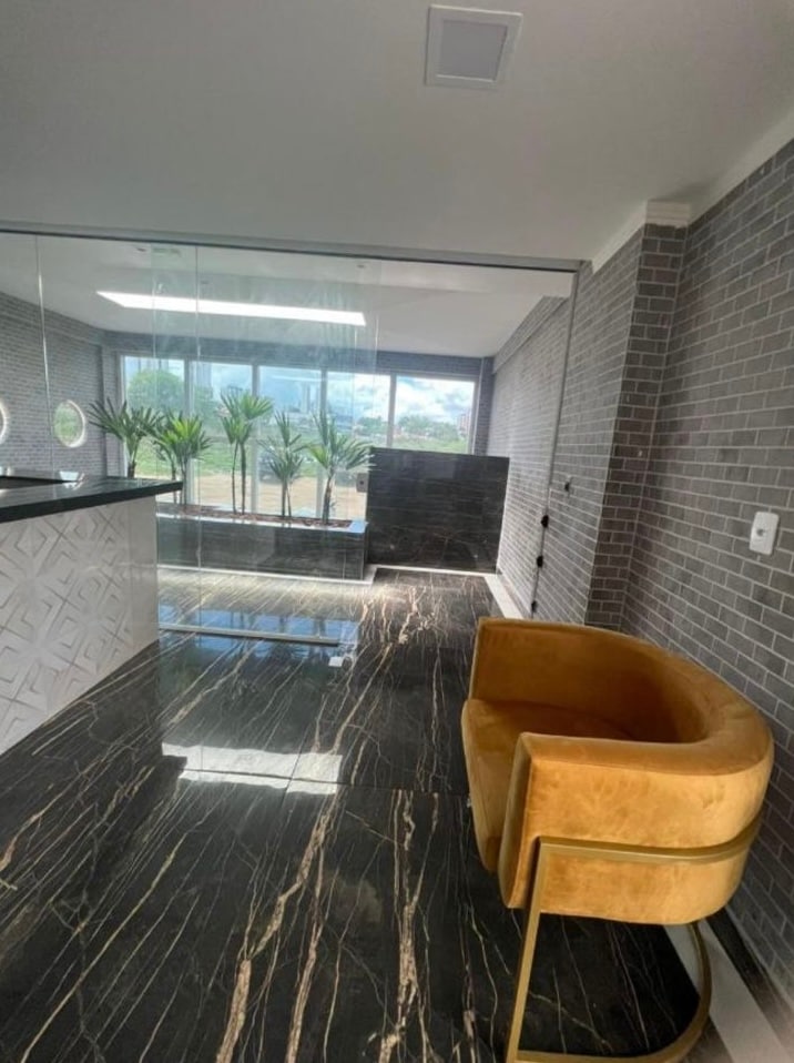 A sleek lobby area is featured with polished black marble flooring, accented by large windows letting in natural light. A contemporary gold armchair sits beside a stylish reception desk, enhancing the modern aesthetic. Green plants are displayed outside the windows, adding a touch of nature.
