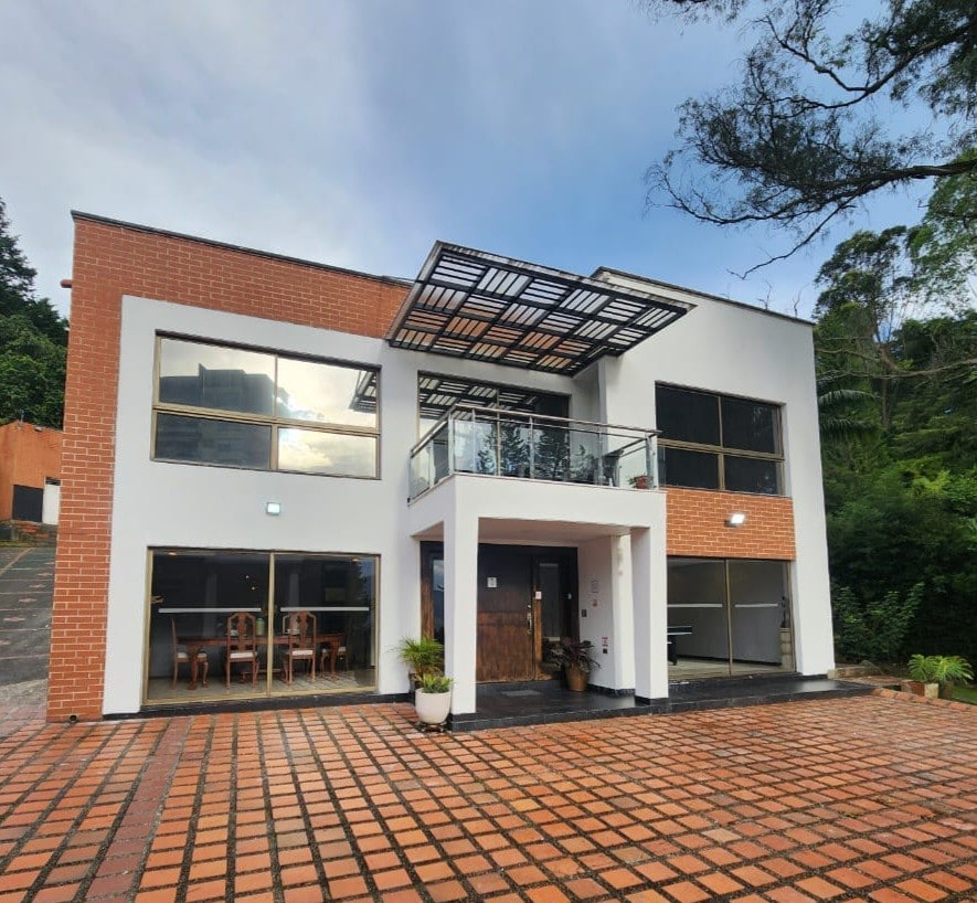 A modern two-story villa features a brick exterior and large glass windows. The welcoming entrance is complemented by a covered balcony extending over the front door. A spacious patio with patterned tiles provides an inviting area for relaxation or gatherings.