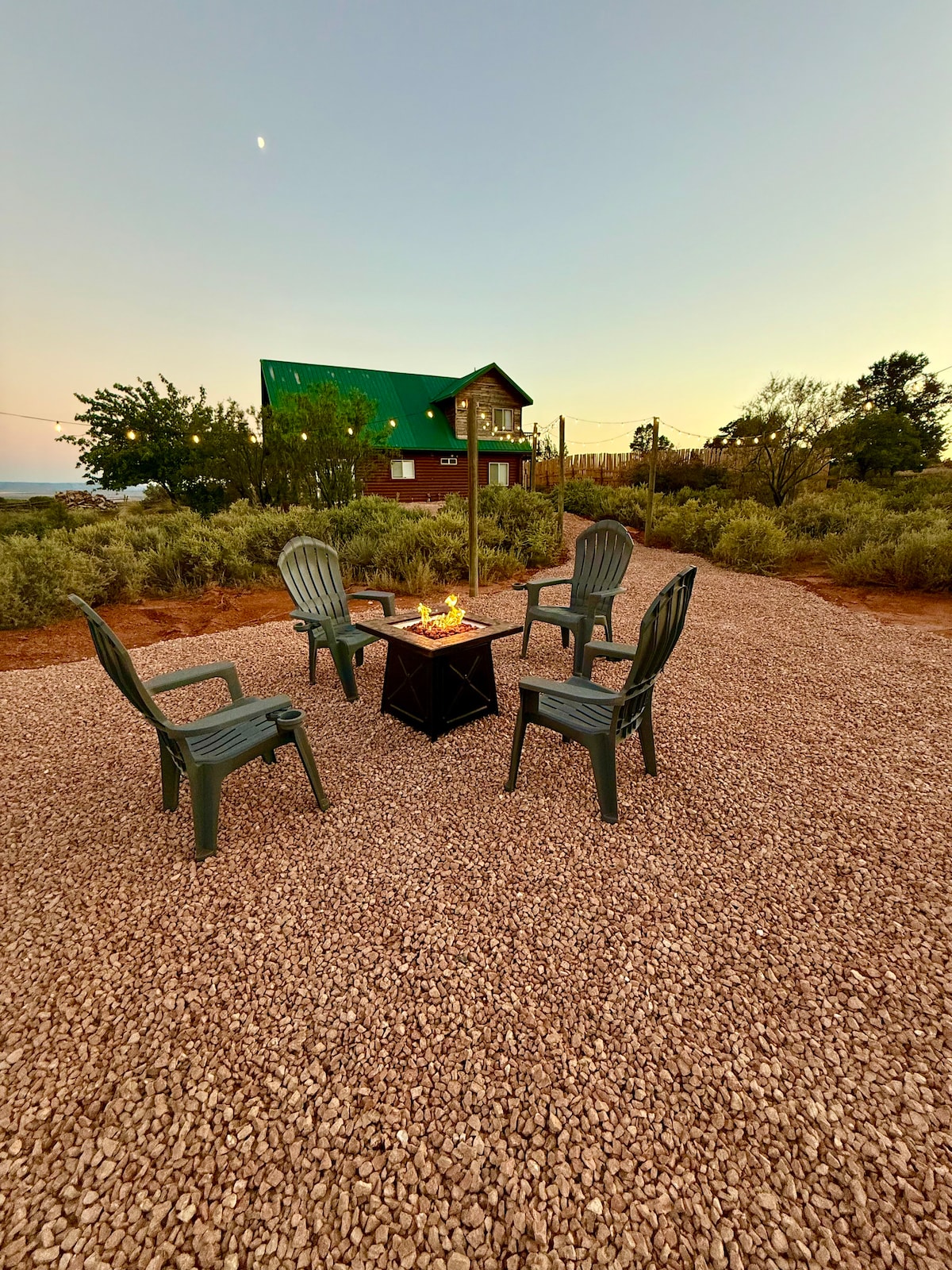 A fire pit area is surrounded by four plastic chairs arranged around a central table. The gravel underfoot is visible, and distant views of a two-story building with a green roof can be seen. Vegetation surrounds the space, providing a natural setting.