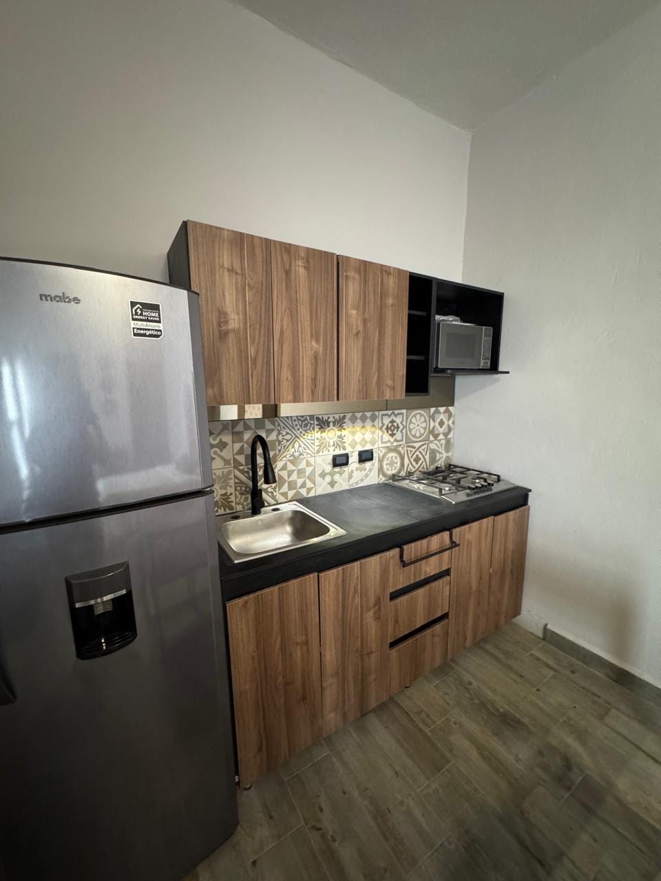 A compact kitchen features wood cabinetry and modern appliances, including a refrigerator and a small microwave. An integrated sink is positioned beneath a stylish backsplash with intricate tile patterns, while a gas stove is set on the counter, completing this functional cooking space.