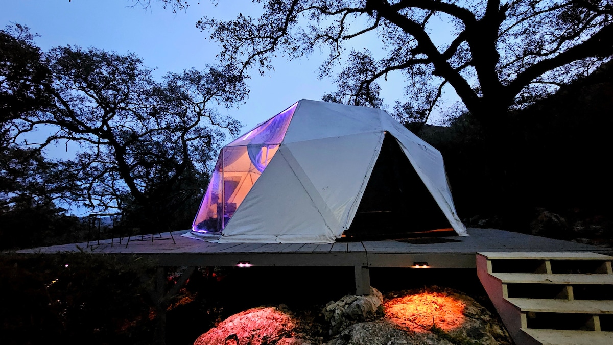 A glamping dome sits amid trees, illuminated with soft lighting from within. The structure's clear panels offer a glimpse of the cozy interior. A wooden deck extends out, enhancing the connection with nature, while the surrounding terrain includes rocky elements and natural vegetation.