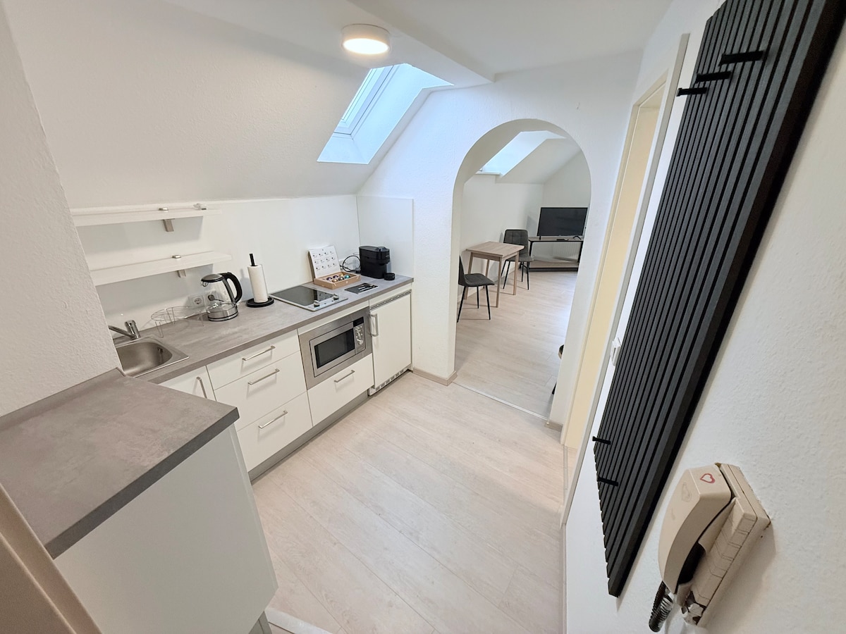 A modern kitchen is visible, featuring white cabinetry and sleek appliances. A small countertop separates the space from the living area, where an archway leads to a dining table and a television. Natural light comes from the skylight overhead, brightening the neutral tones.