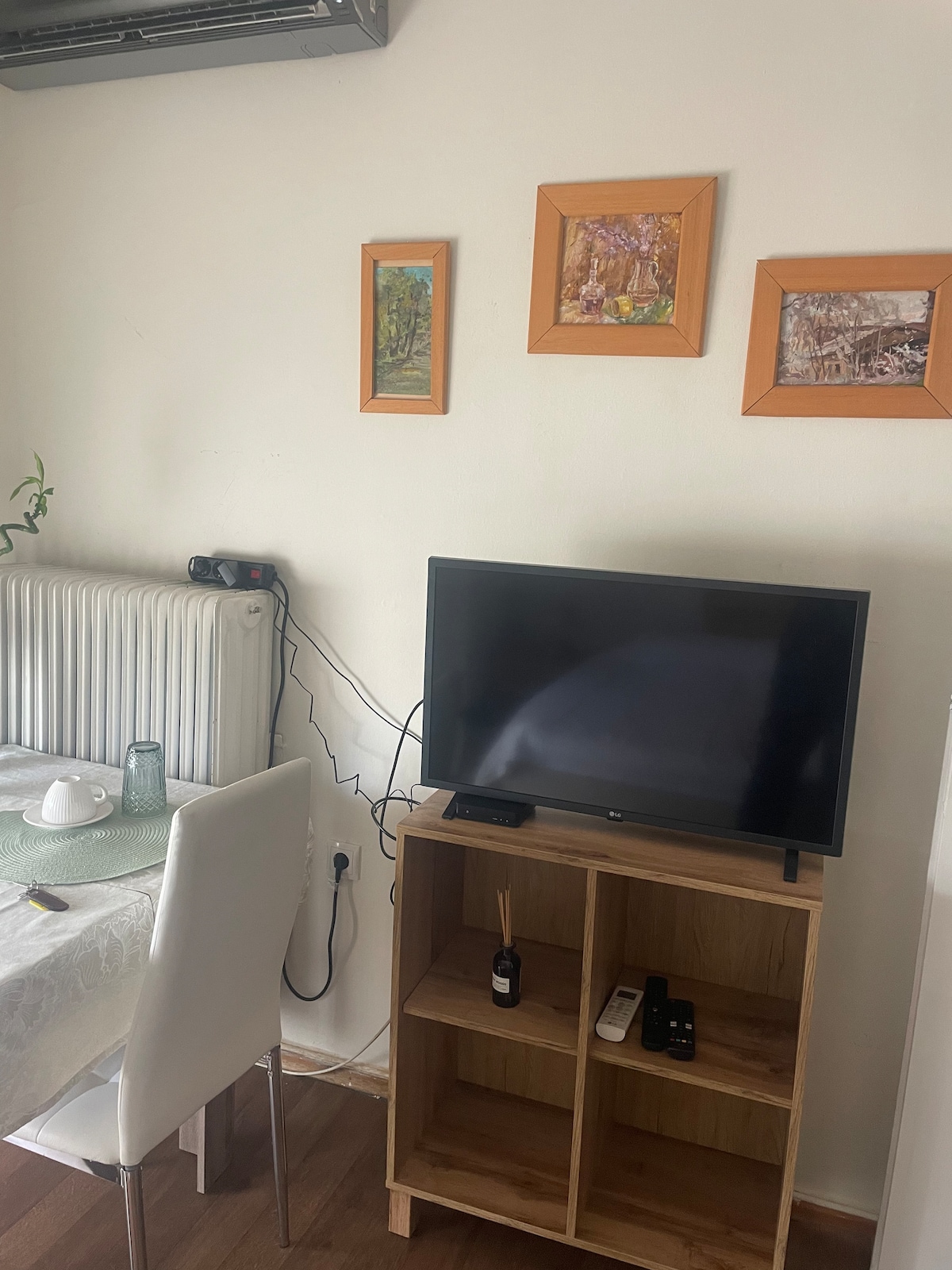 A dining area features a white table set with a teapot and glass. A wooden shelf houses a television and decorative items. Above, three framed artworks display nature scenes. The wall is complemented by a radiator, creating a cozy and functional space.