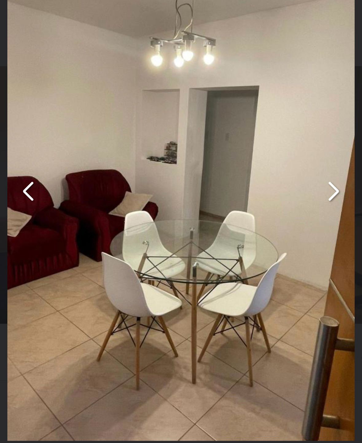 A dining area features a round glass table surrounded by four white chairs with wooden legs. Two red armchairs are positioned nearby, creating a cozy seating arrangement. A simple light fixture hangs above, illuminating the neutral-toned walls and floor tiles.