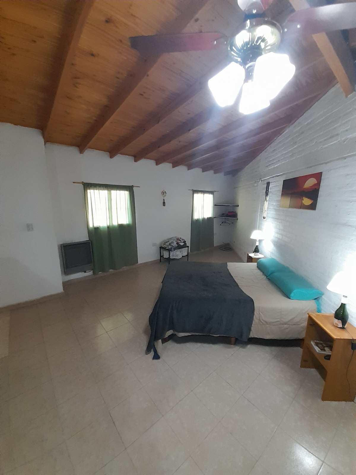 A spacious bedroom features high wooden ceilings and a harmonious color palette. A large bed is positioned against a textured wall, complemented by a bedside table. Natural light enters through two windows dressed with sheer curtains, creating a bright and airy atmosphere.