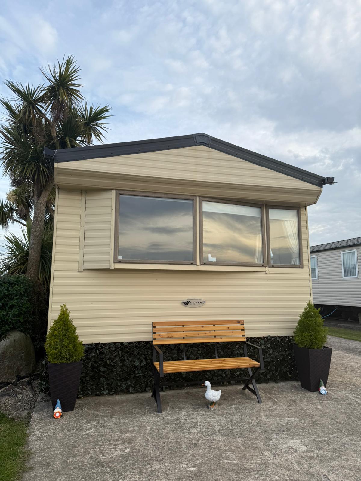 The exterior of the holiday home displays a well-maintained facade with large windows and a welcoming entrance. A wooden bench is positioned in front, flanked by small decorative plants. The scene is set against a backdrop of palm trees and neighboring properties.