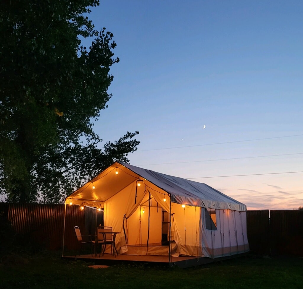 A spacious glamping tent is illuminated by warm lights, set against a twilight sky. A small table and chairs are positioned on the porch, offering a cozy outdoor seating area. Nearby trees add a natural feel to the secluded setting.