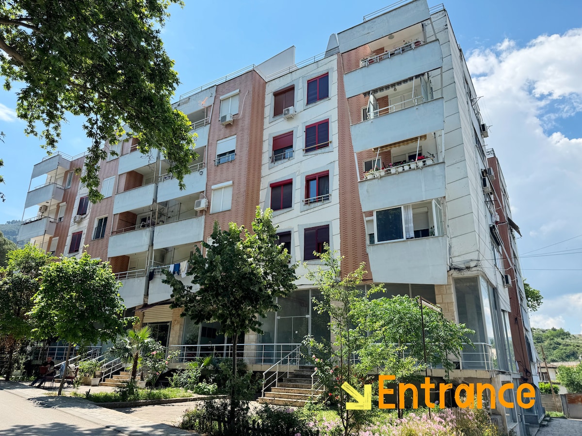 The exterior of a multi-story building displays a combination of beige and white panels. Lush greenery and small trees surround the entrance, enhancing the inviting appearance. Large windows allow natural light into the apartments, while balconies provide additional outdoor space.
