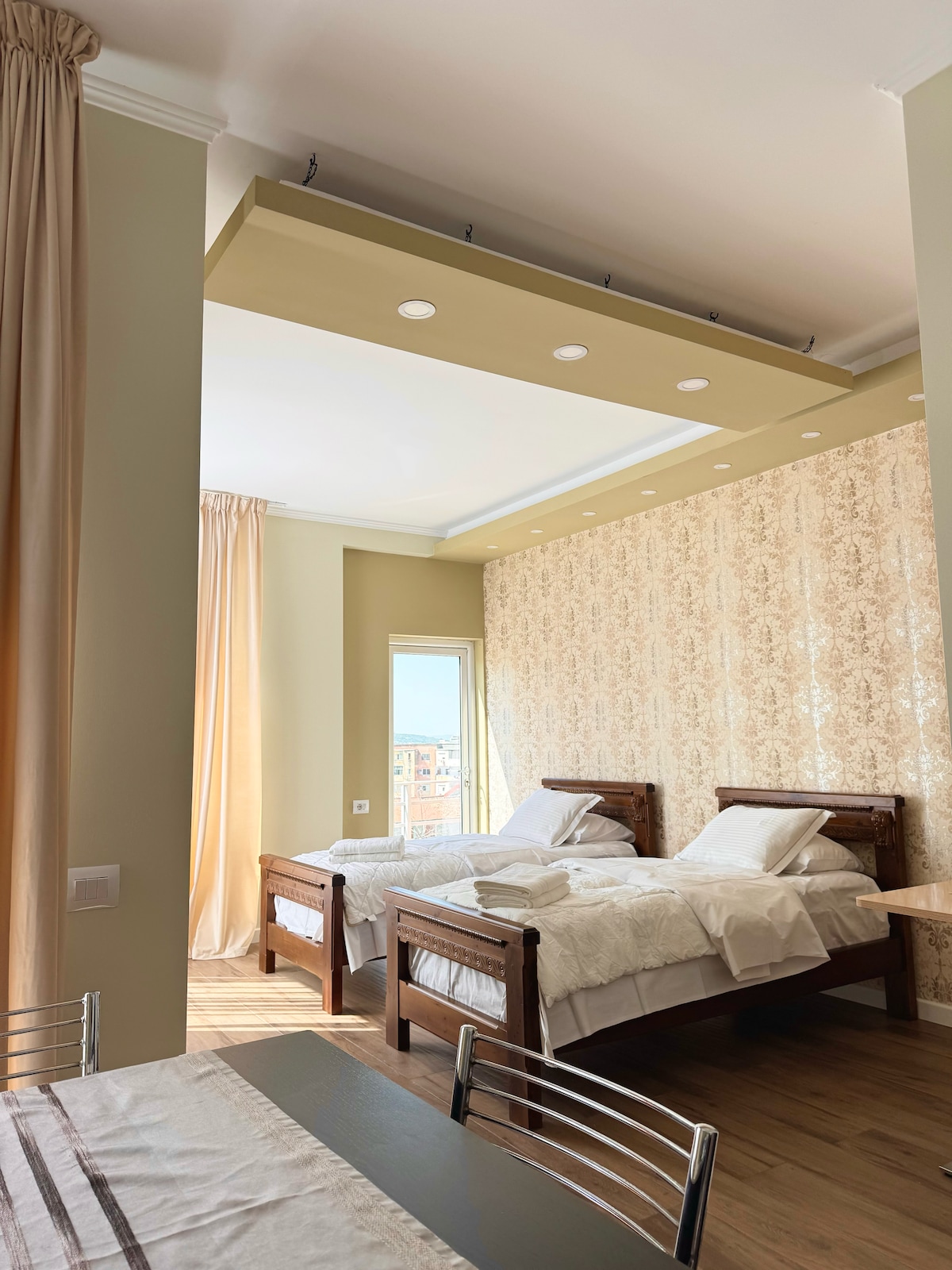 A bright bedroom features two wooden beds with neatly arranged white linens. A soft, warm light is provided by recessed lighting and natural sunlight streaming through a window. Elegant curtains frame the window, and a textured wallpaper adds a subtle design element to the room.