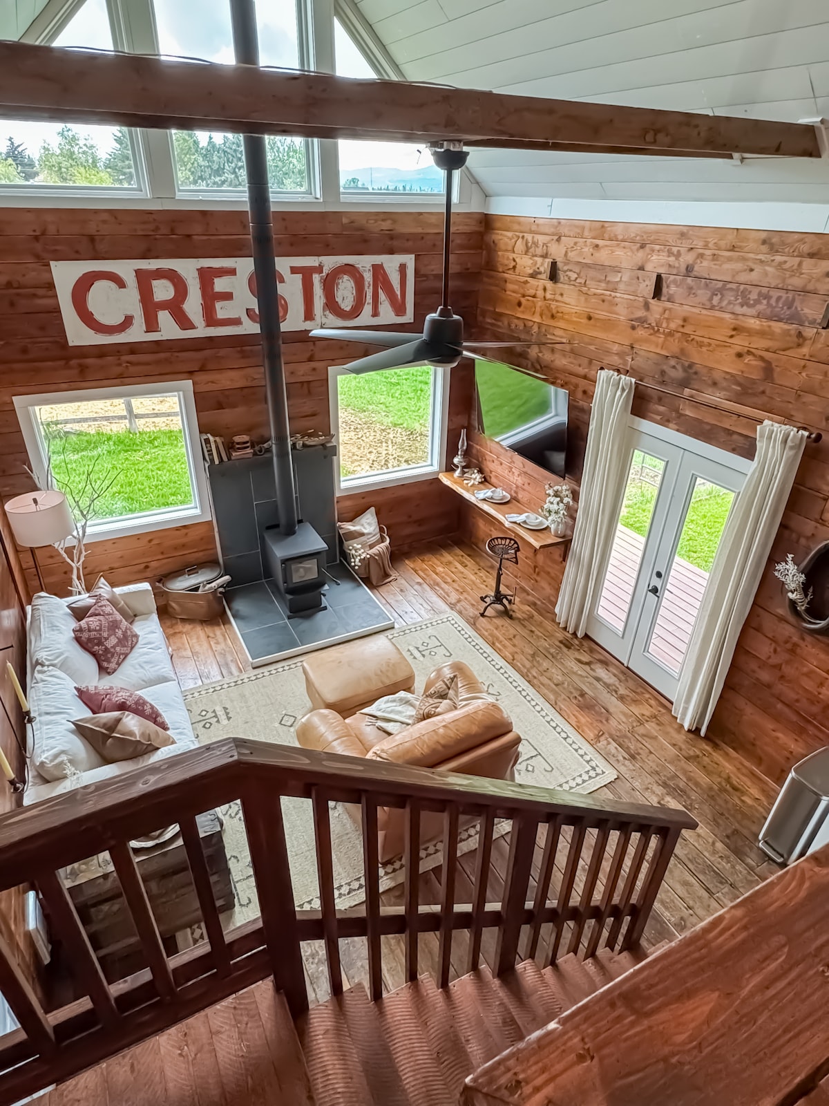 The living area features wooden walls and vaulted ceilings, creating an open and inviting space. A comfortable sofa and armchair are positioned around a central rug. A wood stove provides warmth, while large windows invite natural light and views of the outside.
