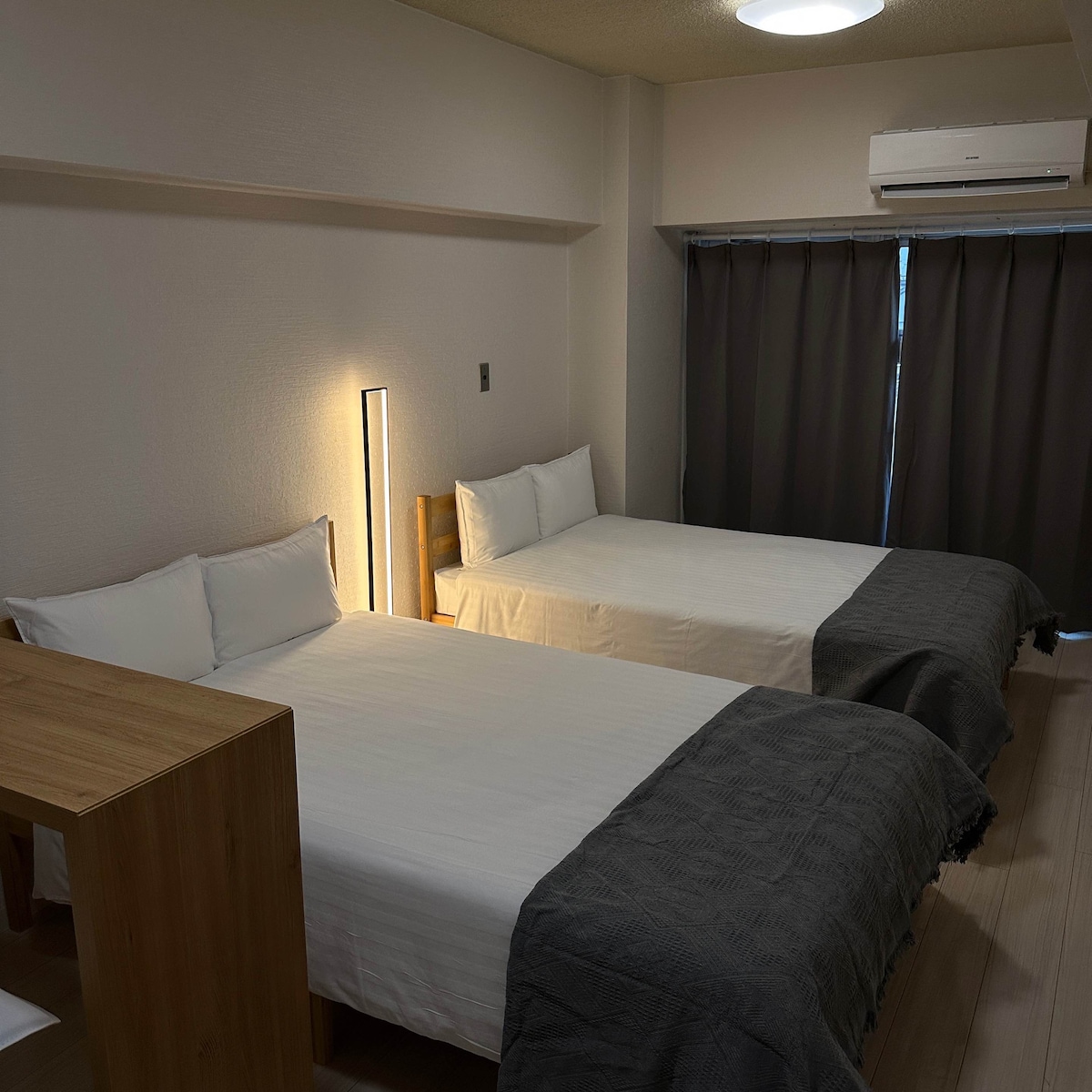 The cozy sleeping area features two spacious beds, neatly made with white linens and grey throws. A bedside lamp adds gentle illumination. A simple wooden table is positioned nearby, while large curtains partially cover a window, contributing to a calm and inviting environment.