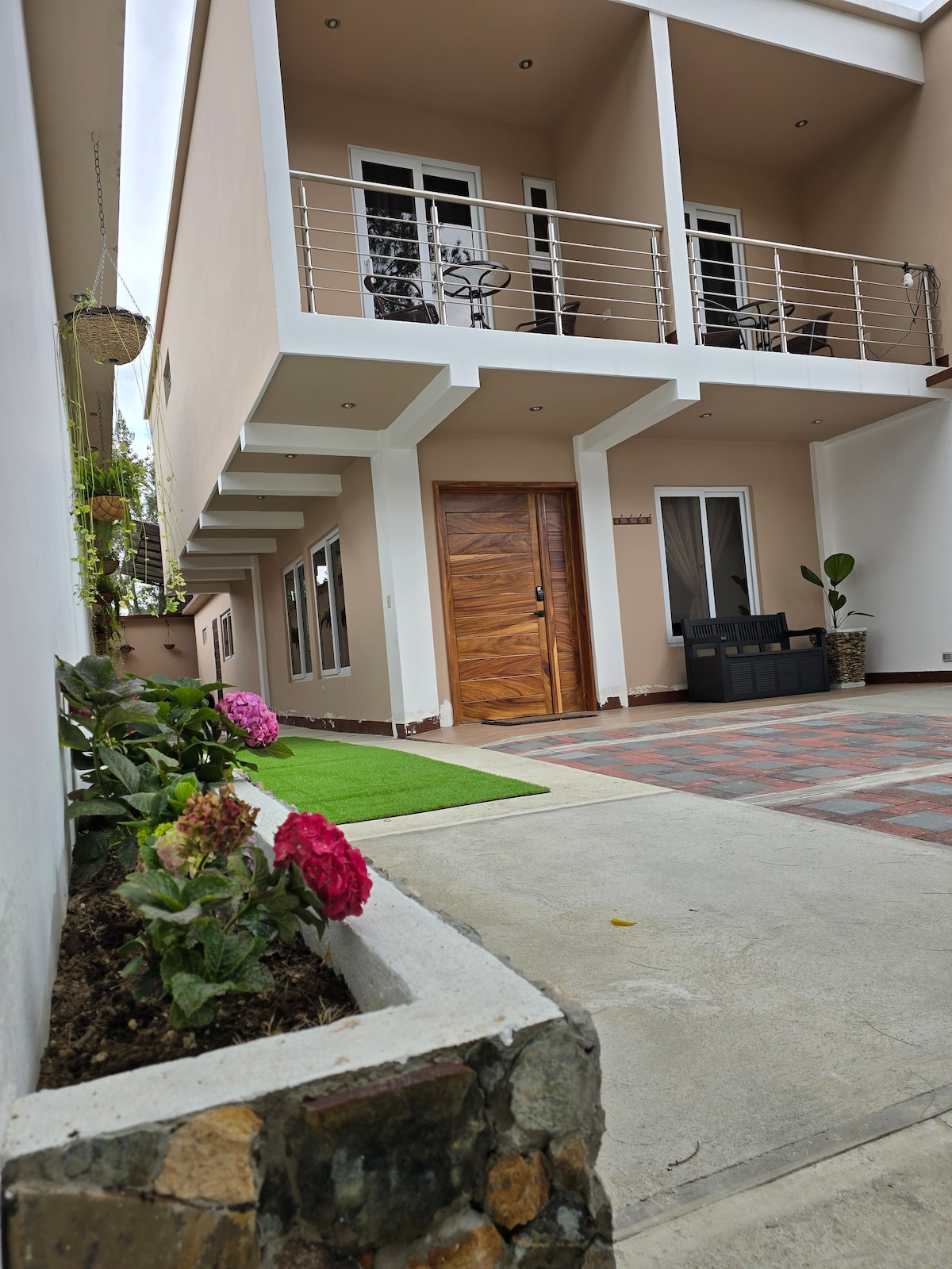 The image shows the exterior of a two-story house with soft pink walls and a wooden front door. A small seating area is visible on the left, while vibrant flowers are planted along the side, enhancing the welcoming environment. A paved driveway leads to the entrance.