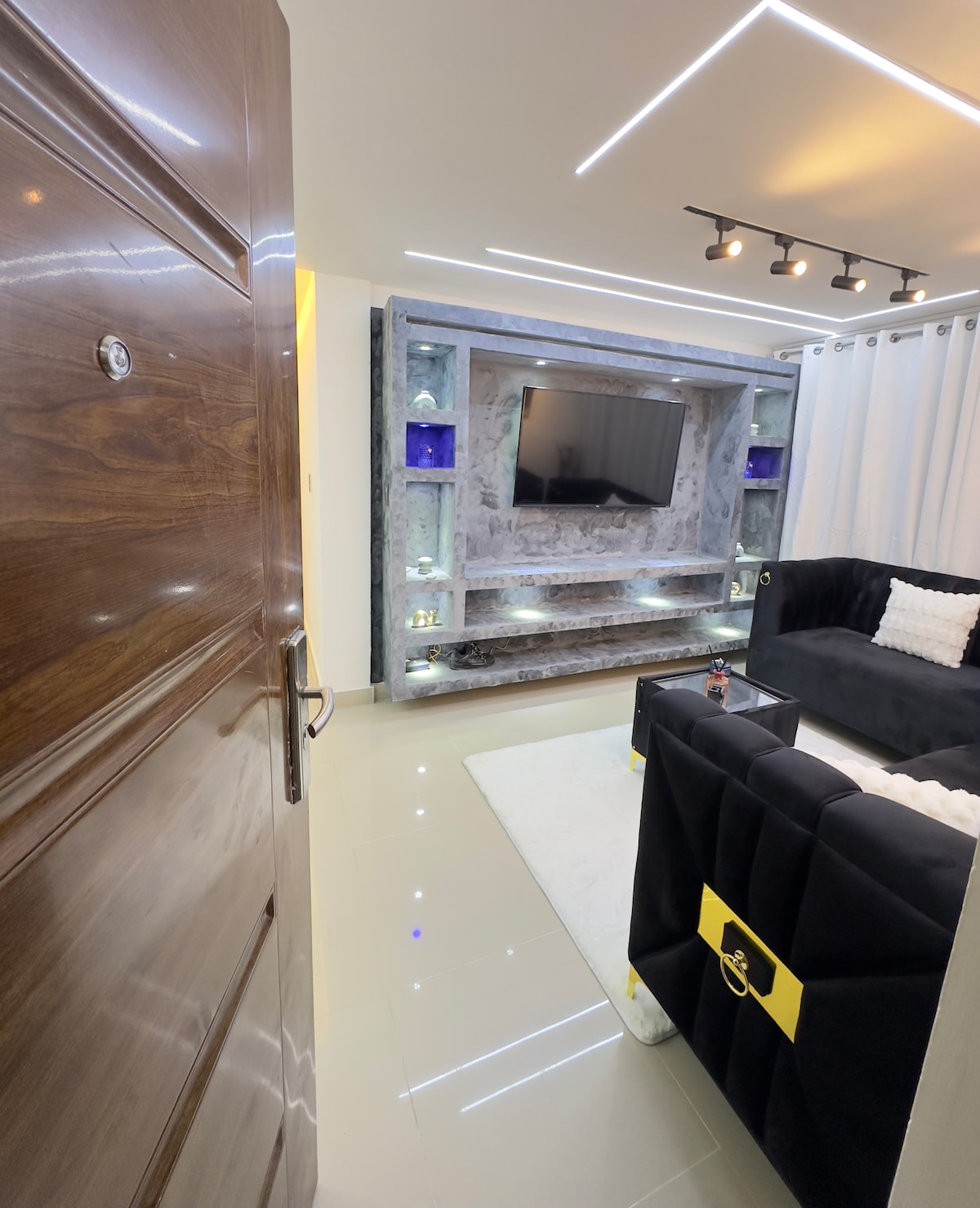 A modern living area is visible upon entry, featuring a sleek black sofa and a coffee table. A wall-mounted flat-screen TV is set against a stylish backdrop with ambient lighting. Soft white curtains frame a large window, enhancing the sense of space and light.