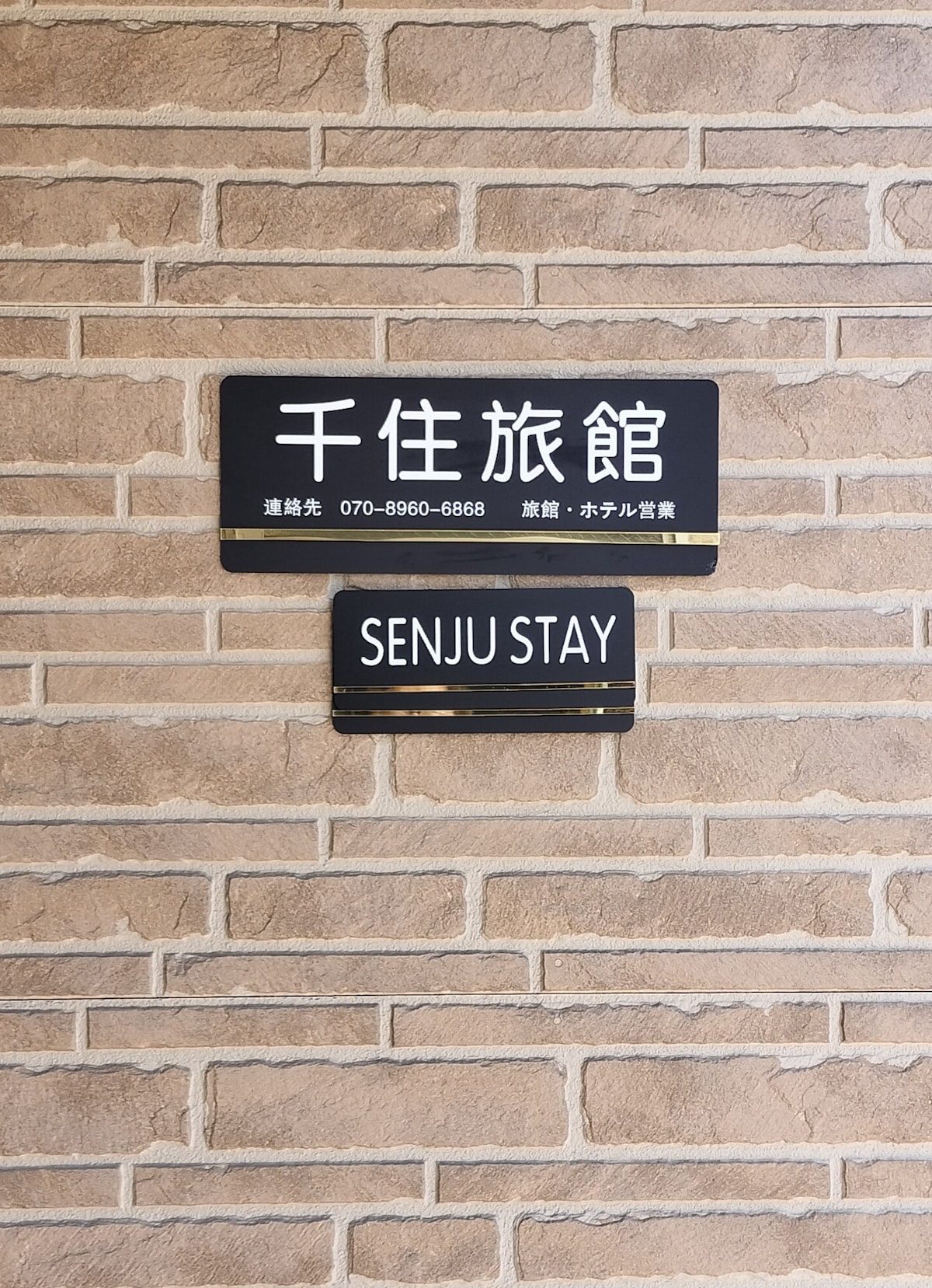 The entrance features a rustic brick facade with two signs. The top sign displays the name in both Japanese and English, identifying the property as 'Senju Stay.' The signage is clear and legible, contributing to the welcoming ambiance.