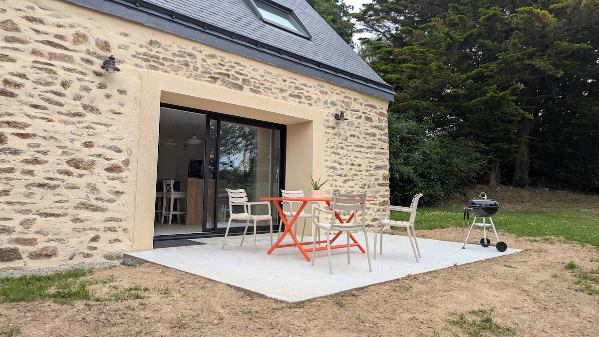 An outdoor terrace features a dining set, consisting of a round table and four light-colored chairs. A charcoal grill is positioned nearby, with a backdrop of greenery from surrounding trees. The terrace is part of a stone-clad house with large glass doors leading to the interior.