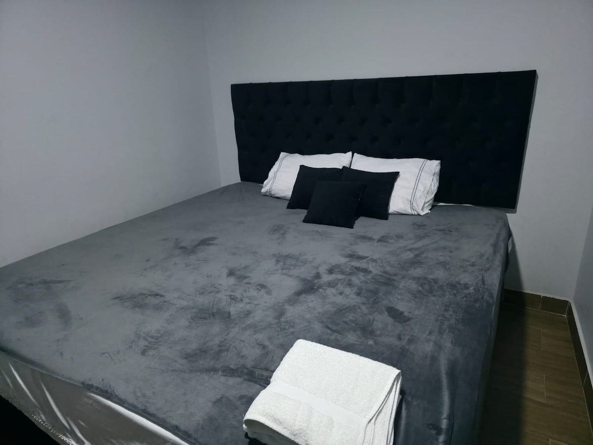 A spacious bed with a plush gray blanket and black tufted headboard is set against a soft gray wall. Two decorative pillows enhance the bed's comfort. A fresh white towel is neatly arranged on the side, providing a welcoming touch to the sleeping area.