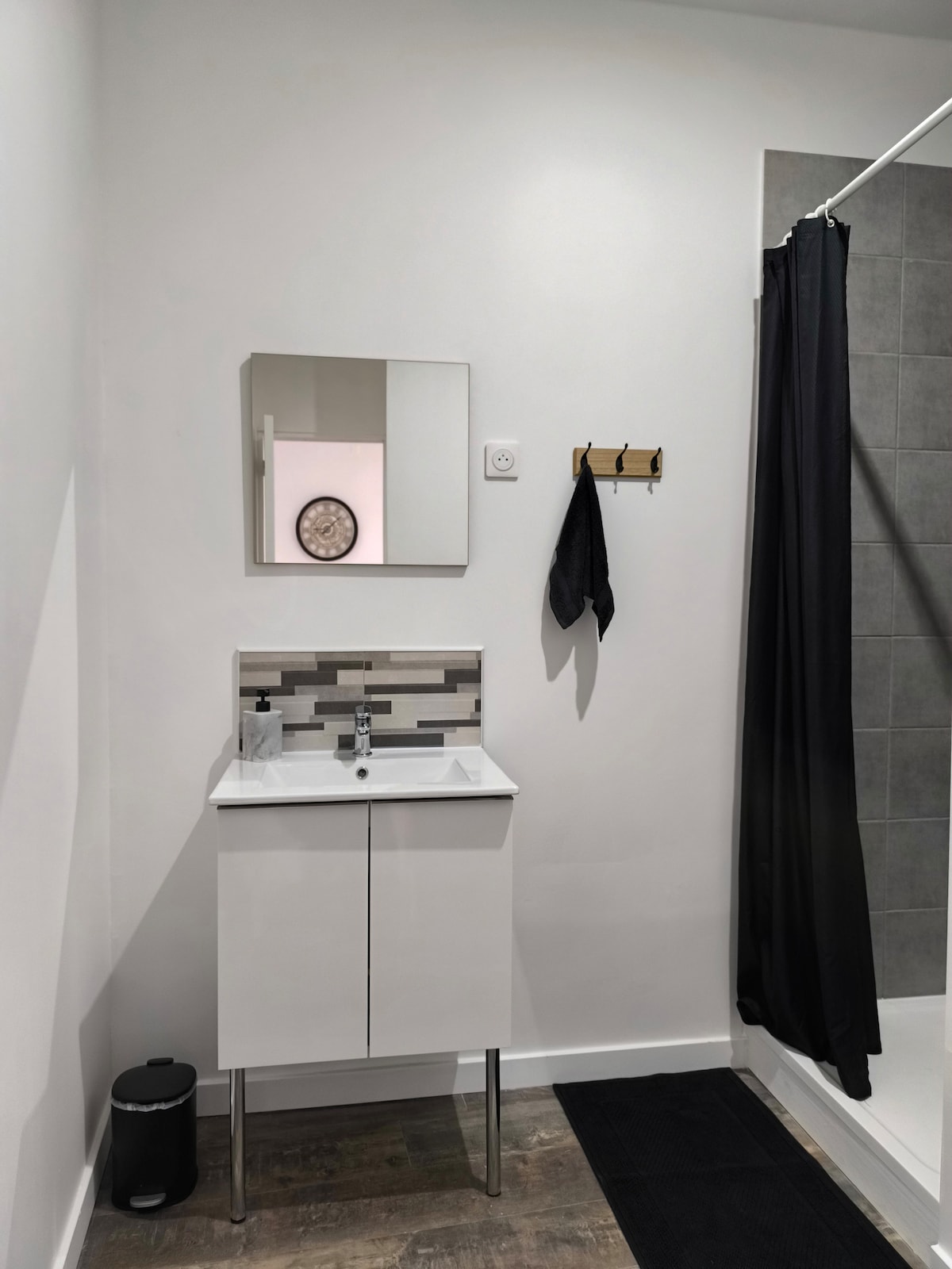 A modern bathroom features a compact vanity with a rectangular mirror above it. A patterned backsplash complements the clean white cabinetry. A shower area is visible on one side, with a dark curtain and textured gray tiles. A towel hangs on a wall hook nearby, and flooring is wood-like.