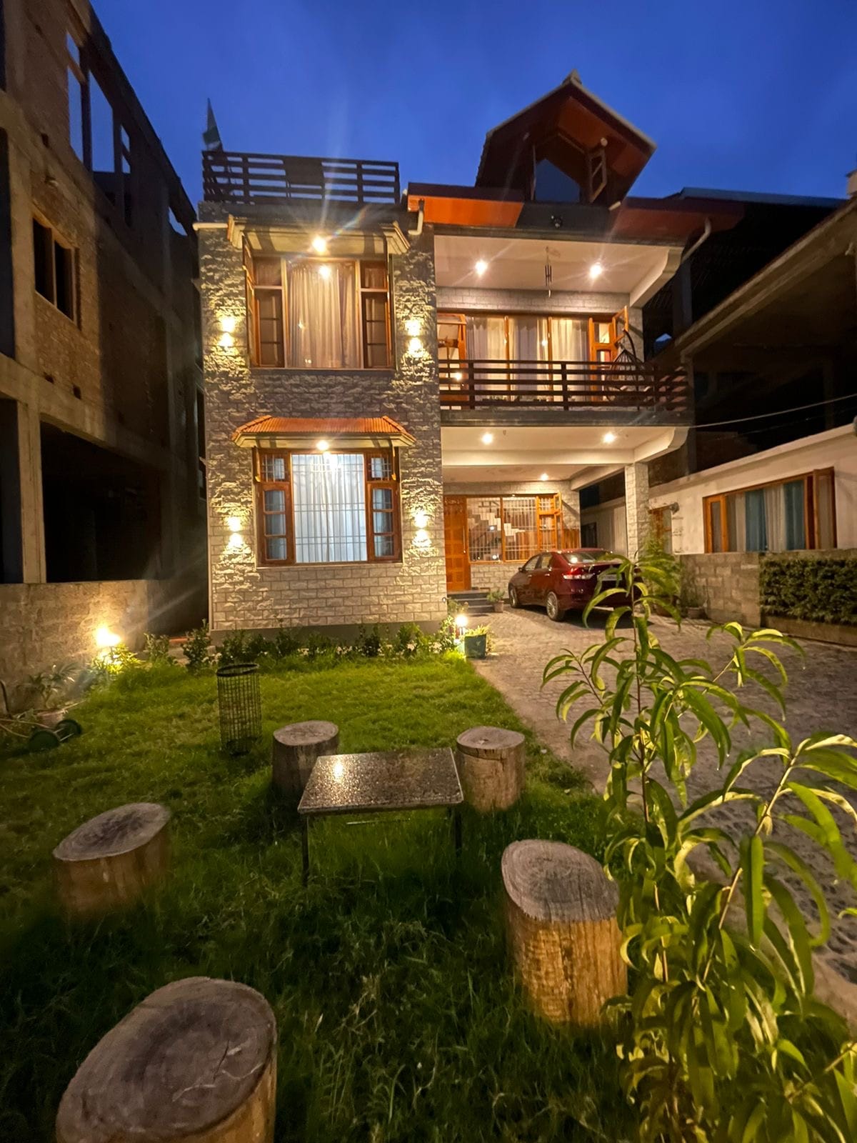 A two-story house is illuminated at dusk, with warm lights highlighting its stone exterior. A well-kept garden features a grassy area and round wooden stools arranged around a small table. A vehicle is parked in front, and balconies add to the structure's charm.