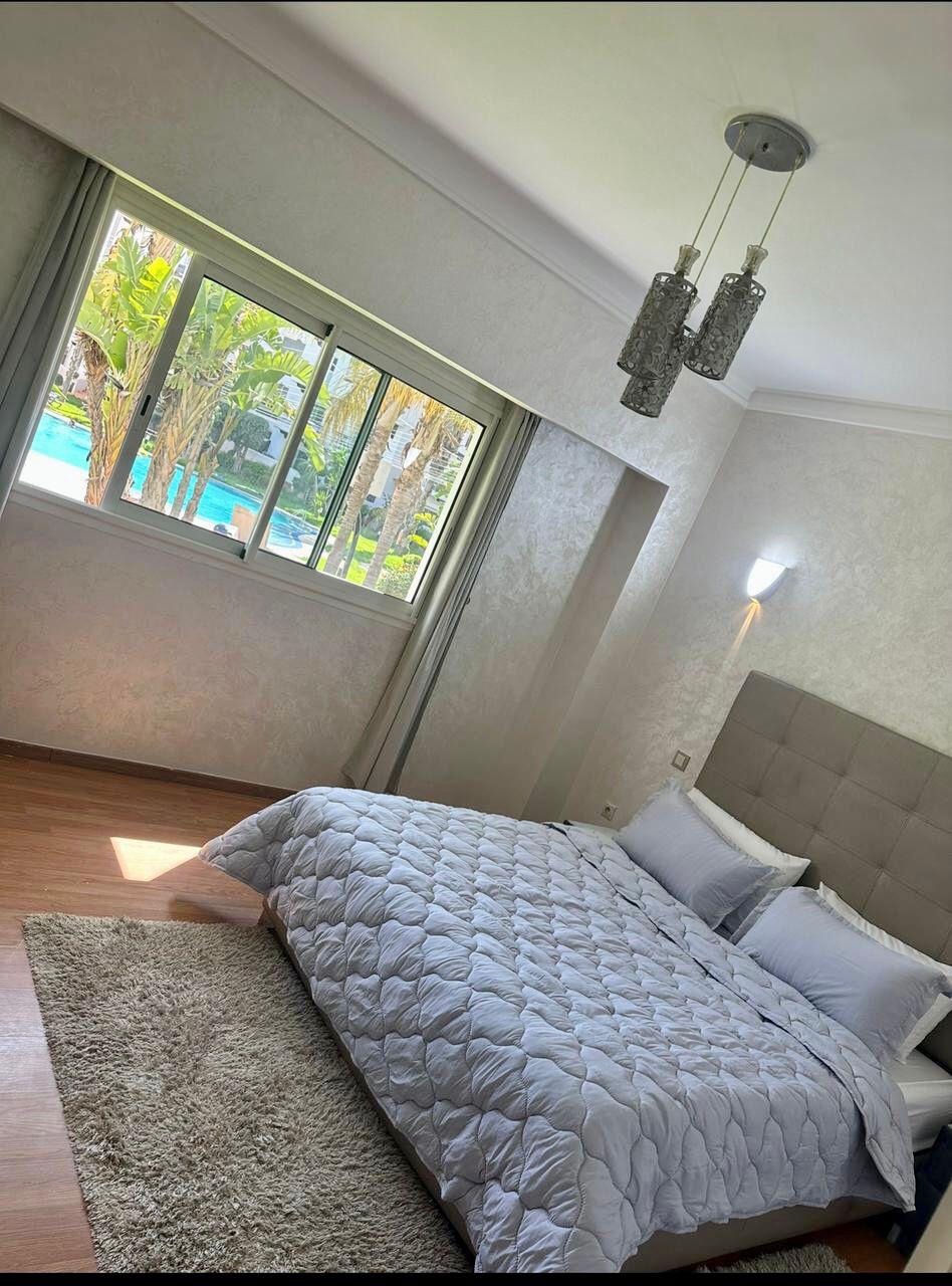 A bright sleeping area features a spacious bed with a soft grey quilt. Natural light streams in through a large window, revealing palm trees and a swimming pool outside. A plush area rug complements the wooden flooring, adding a touch of warmth to the space.