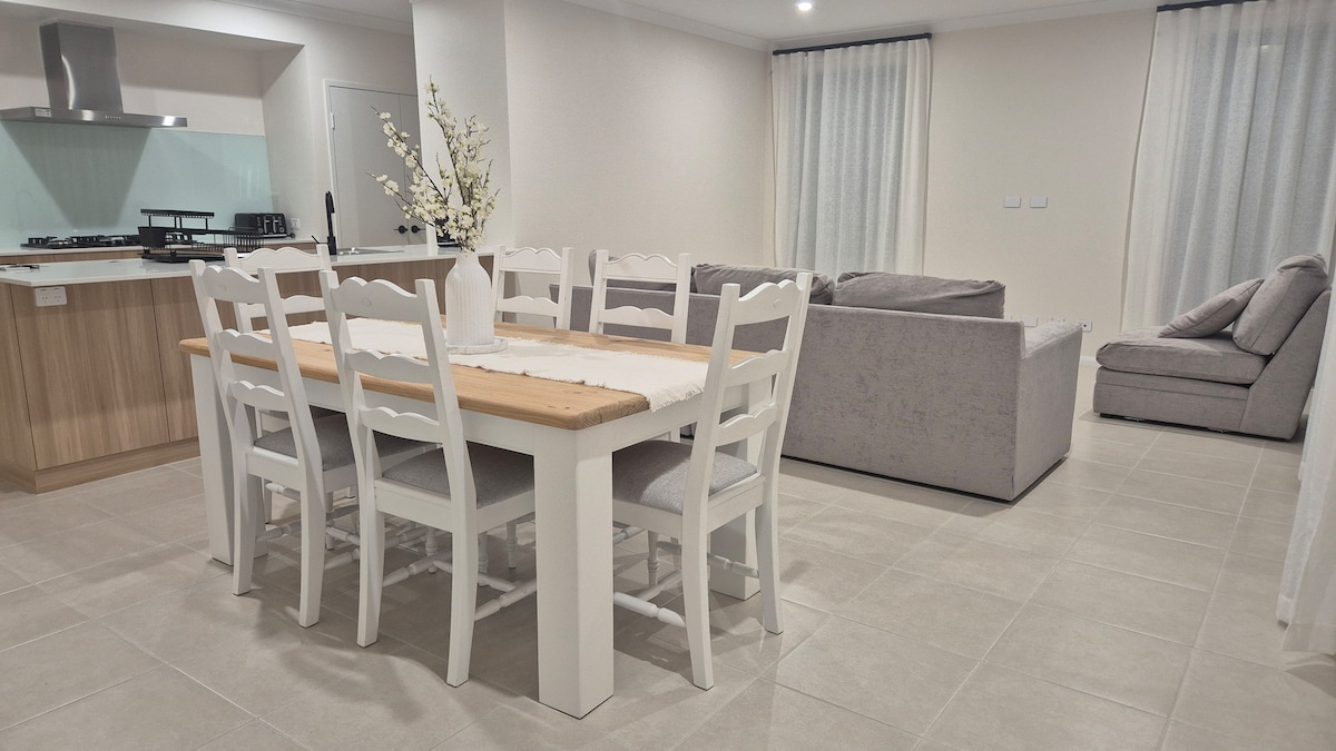 An open living space features a dining table with white chairs and a light wood tabletop. A cozy seating area is visible with a grey sectional sofa and an additional chair. Soft textures and neutral tones create a calm environment, complemented by natural light from large windows.