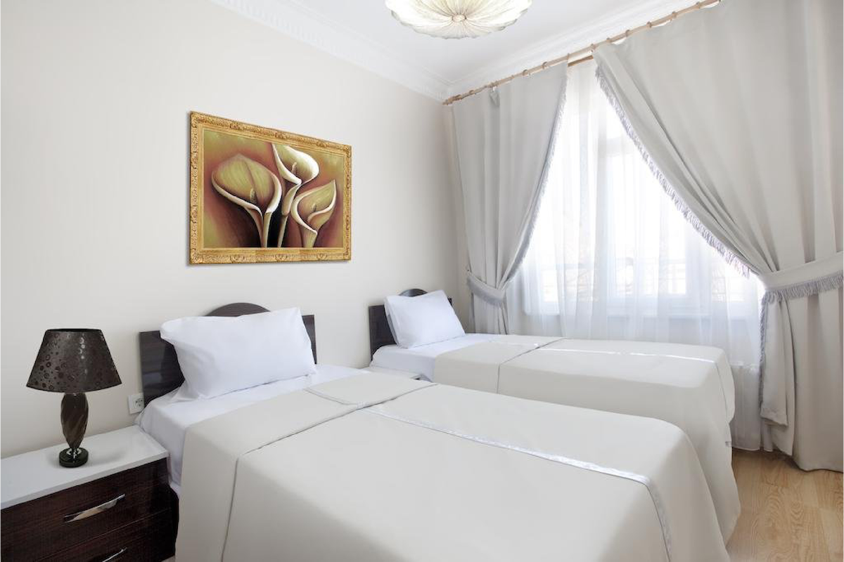 A well-lit room features two beds with white linens, positioned next to dark wooden nightstands. A decorative lamp stands on one side, while a vibrant artwork of flowers adorns the beige wall. Natural light enters through sheer curtains, enhancing the room's serene ambiance.