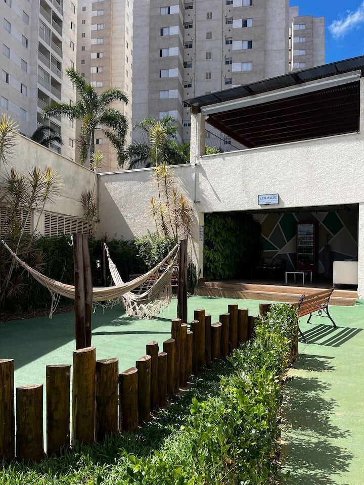 A communal outdoor space is featured, showcasing a hammock gently swaying between wooden posts and a relaxed seating area under a shaded roof. Lush greenery surrounds the area, and tall apartment buildings create a backdrop, contributing to a private and serene atmosphere.