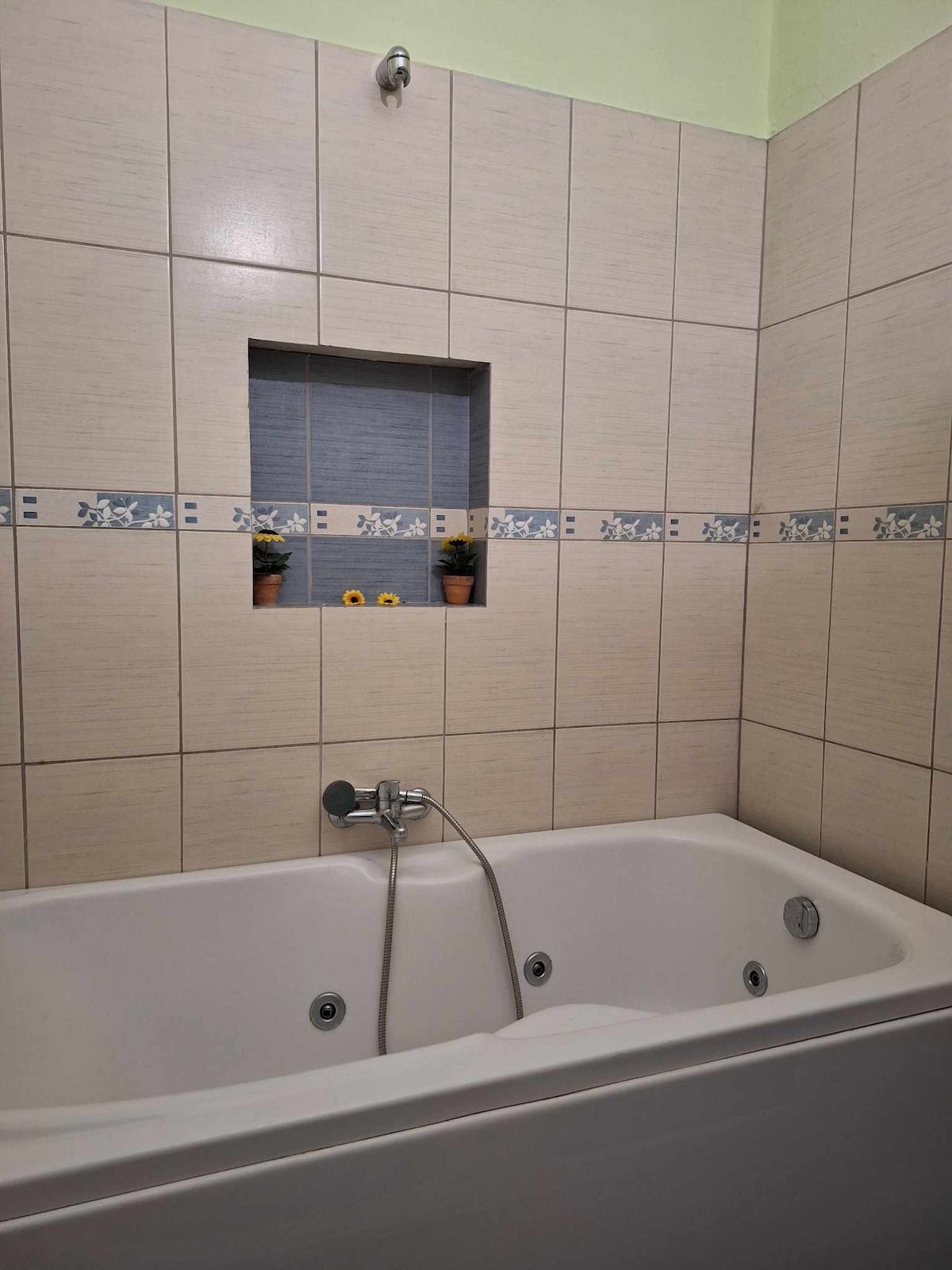 A spacious bathtub is featured against a tiled wall, accented by a decorative niche holding small potted plants. Neutral tiles create a calming atmosphere, while the modern faucet and sleek design contribute to a clean, uncluttered look.
