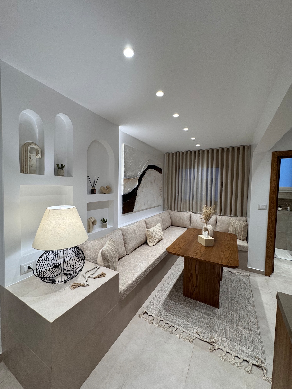 A modern living area features a light-colored sofa arranged against a wall, accompanied by a wooden dining table. Decorative alcoves hold art and small plants. Soft lighting illuminates the space, while a neutral rug adds a cozy touch beneath the table.