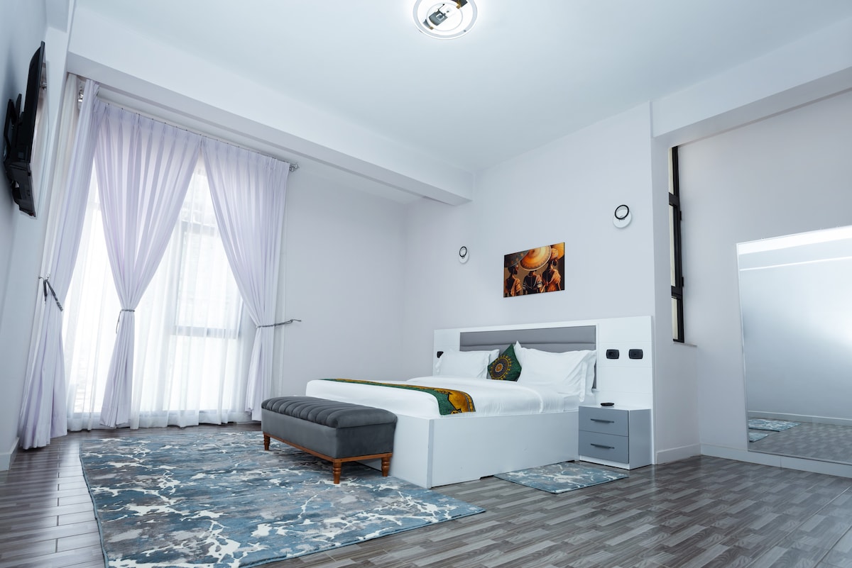 A spacious sleeping area features a large bed with white linens and decorative pillows. A bench is positioned at the foot of the bed. Light-colored curtains frame the window, and a large mirror is visible on one wall, complemented by a decorative rug beneath.