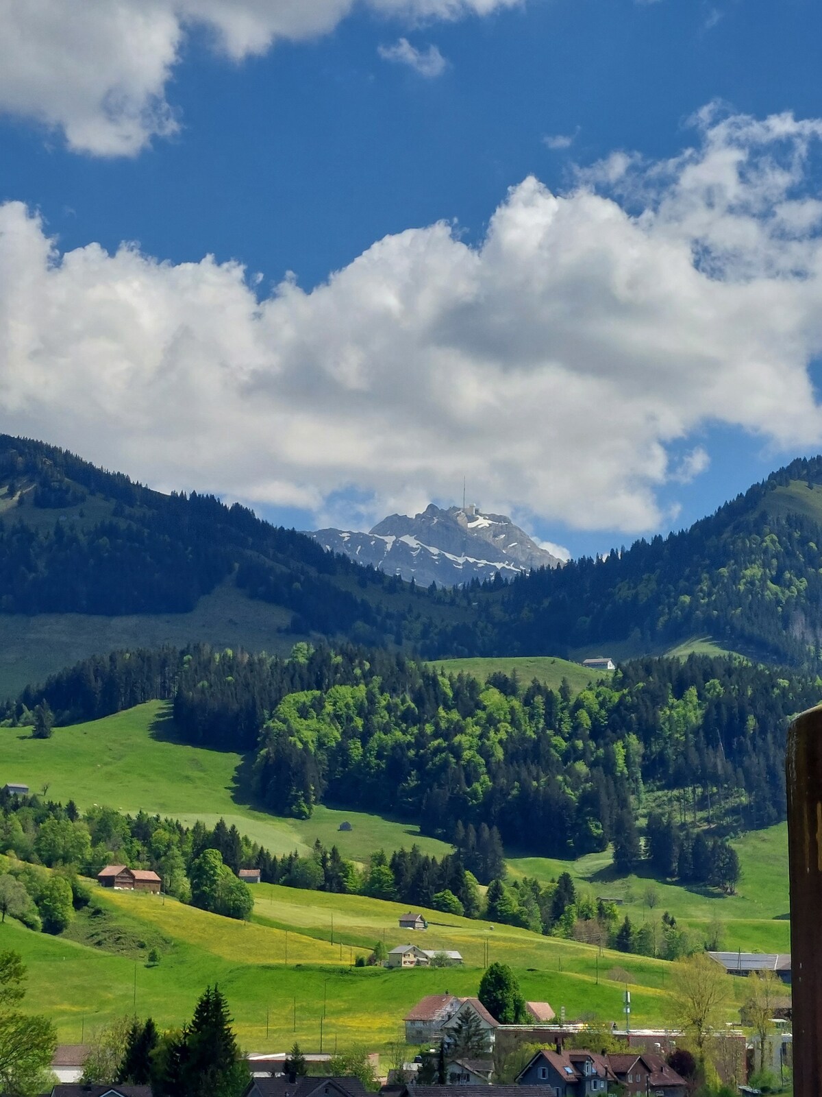 Lush green hills and a vibrant landscape are depicted, with distant mountains capped in snow. Fluffy clouds drift across a bright blue sky, creating a serene natural backdrop. Small villages with scattered houses can be seen nestled in the valley.