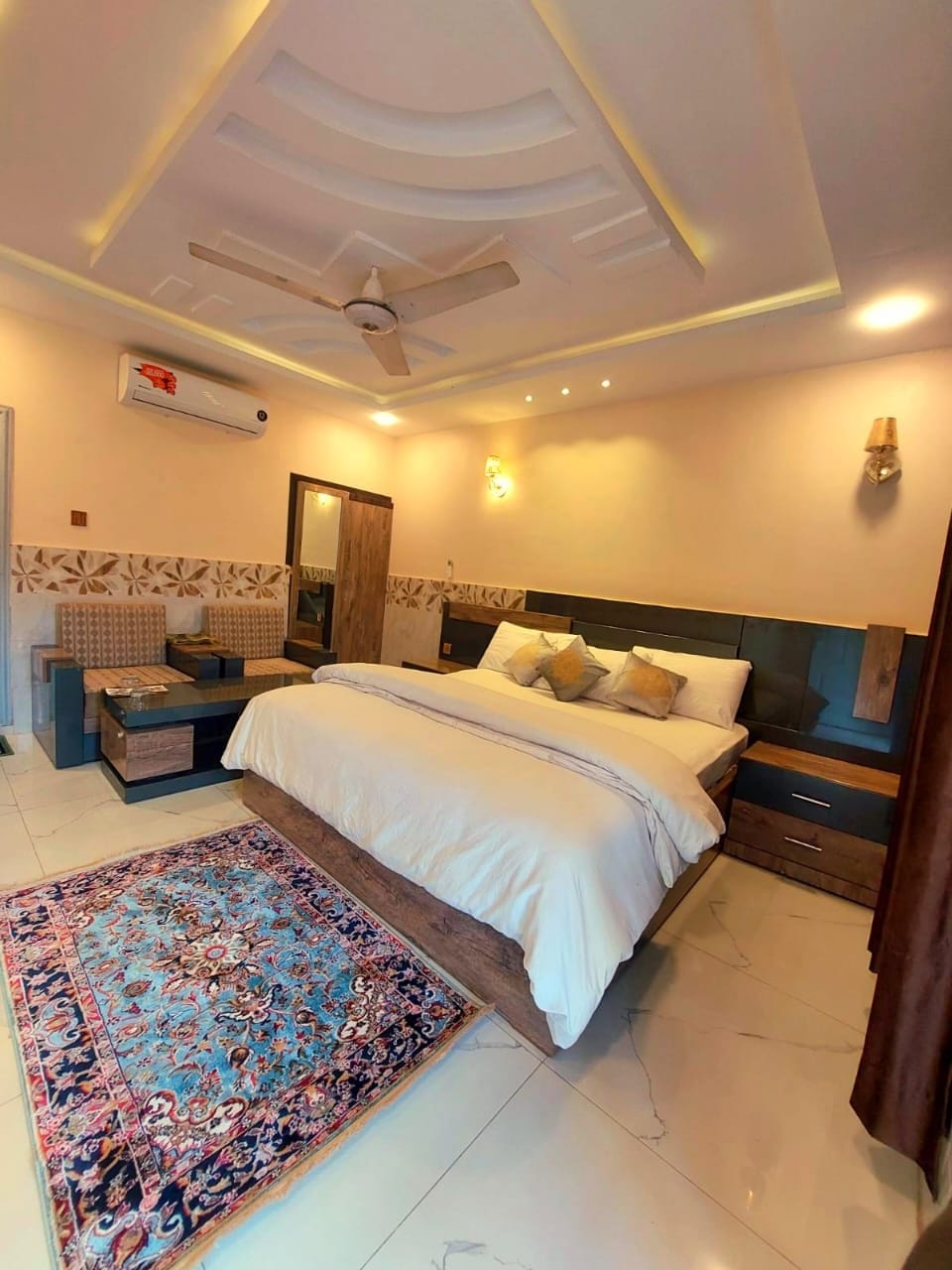 A well-appointed bedroom features a spacious bed with decorative pillows, surrounded by contemporary wooden furniture. Comfortable seating areas are visible, positioned next to a small table. A vibrant area rug adds color, while soft lighting enhances the room's inviting atmosphere.