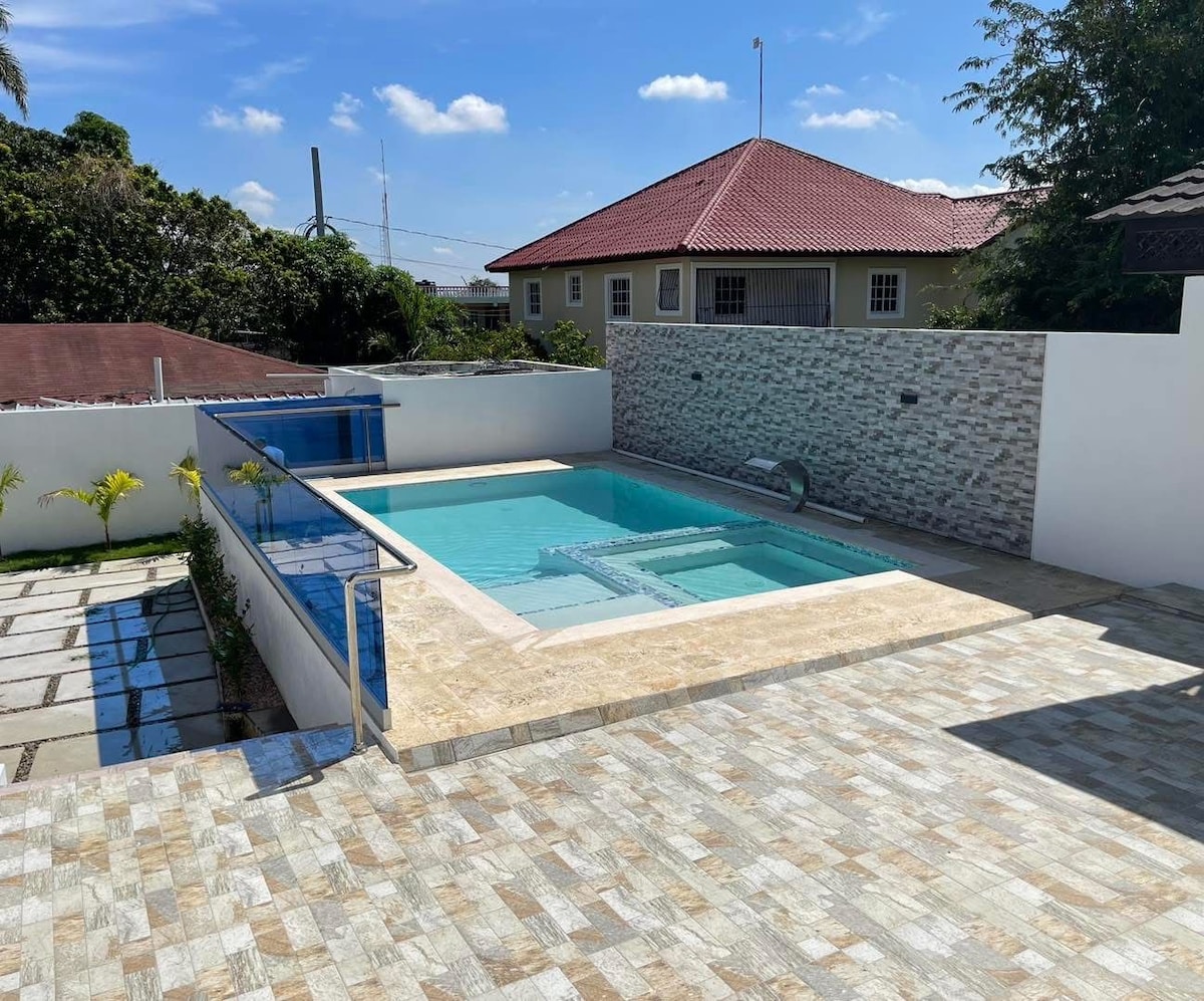 A modern pool area features a clear blue swimming pool enclosed by a glass barrier. Surrounding the pool is a spacious tiled deck, complemented by neatly arranged tropical plants. The background showcases a house with a red roof, enhancing the serene outdoor atmosphere.