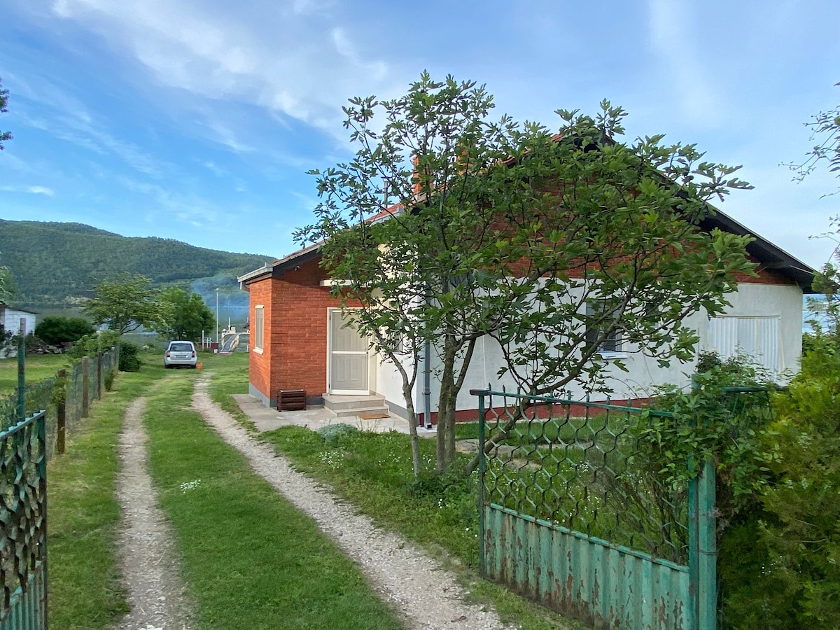 A charming exterior view of a red-brick home, set against a backdrop of rolling hills. Lush greenery surrounds the property, with a tree positioned near the entrance. A winding gravel path leads to the house, providing a welcoming approach for guests arriving by car.