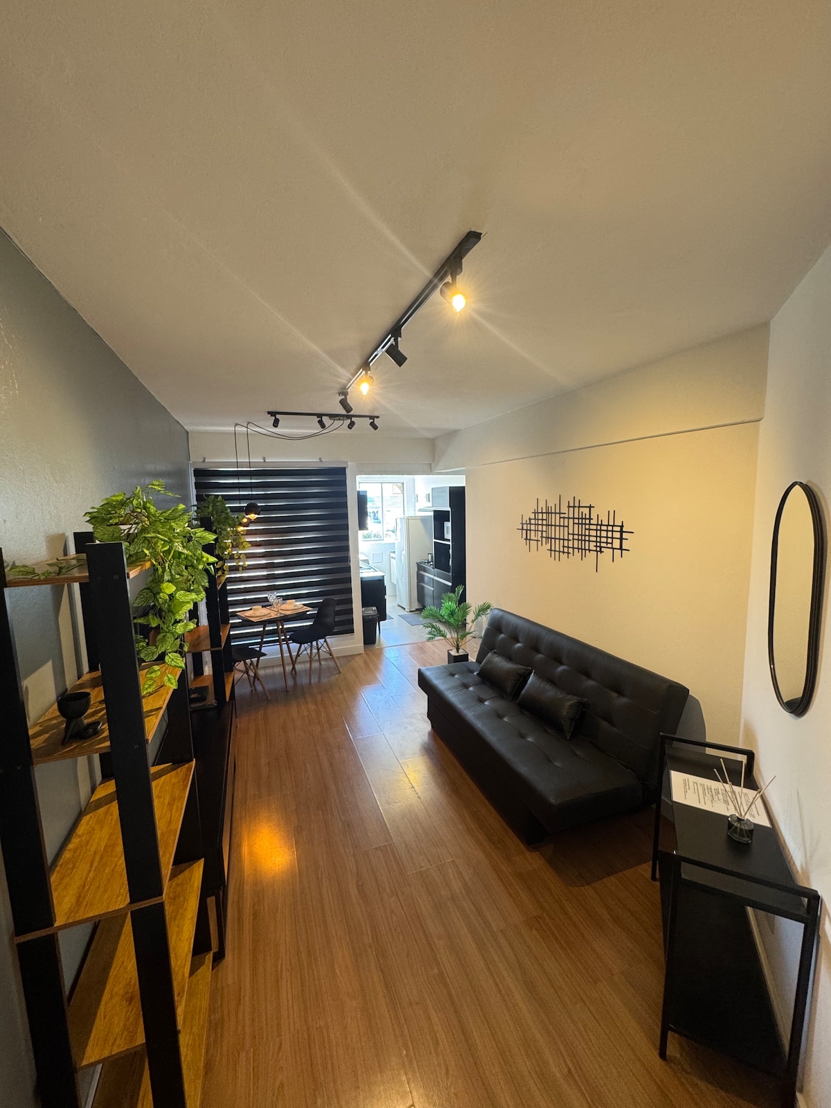 A sleek living area features a modern black sofa positioned across from a minimalist black shelving unit displaying decorative items and greenery. Warm lighting from track fixtures illuminates the space, while a dining table is visible in the background, inviting relaxation and comfort.