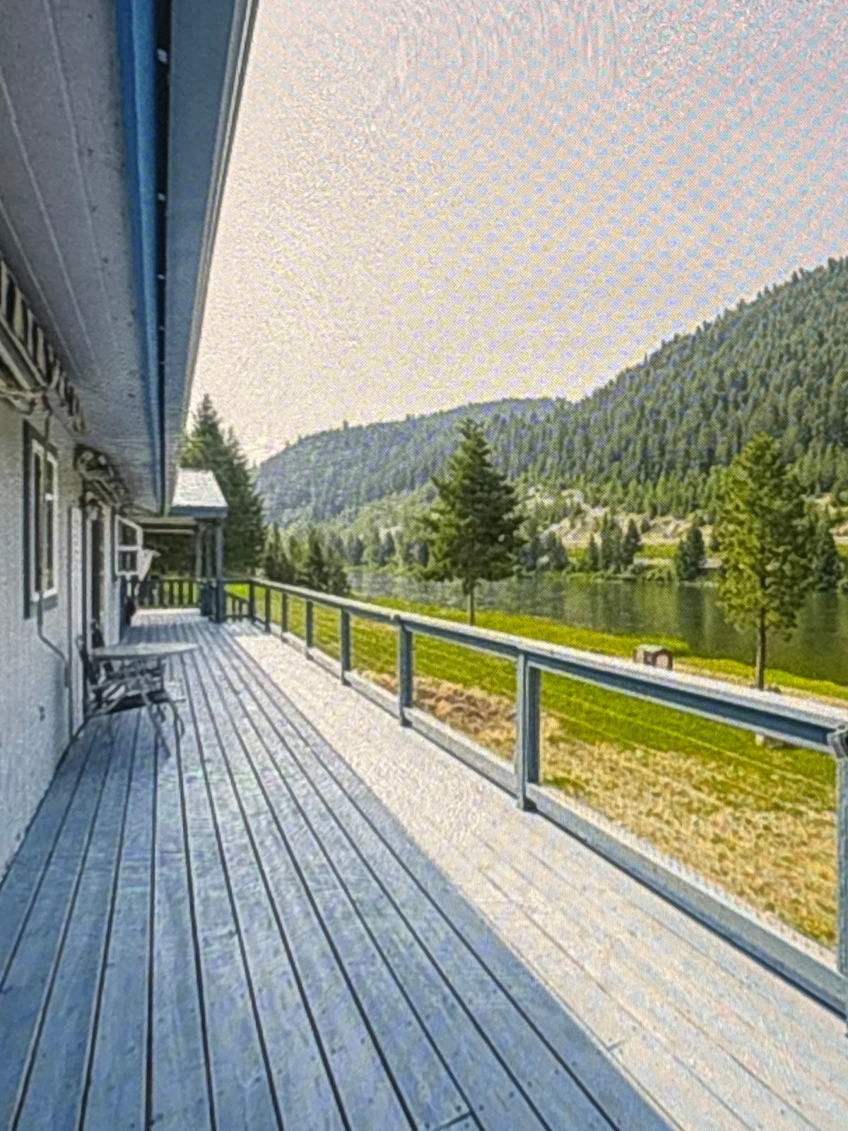 An expansive deck runs along the side of the house, offering views of the serene water and surrounding hills. Tall trees are visible in the distance, and a peaceful green landscape extends toward the water's edge.