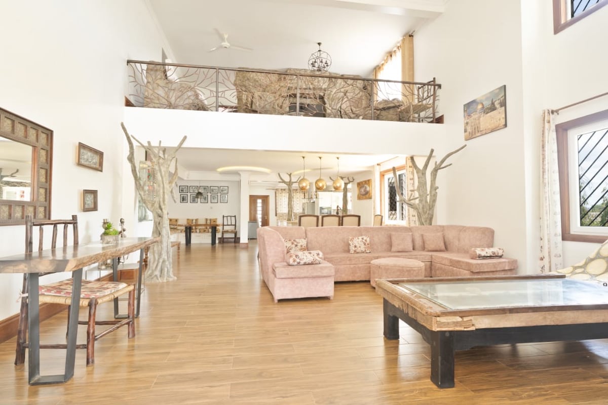 A spacious living area features a large pink sectional sofa and a wooden coffee table with a glass top. Natural light streams through large windows, highlighting the warm wooden flooring. An adjacent dining space includes a wooden table and chairs, with decorative elements enhancing the environment.