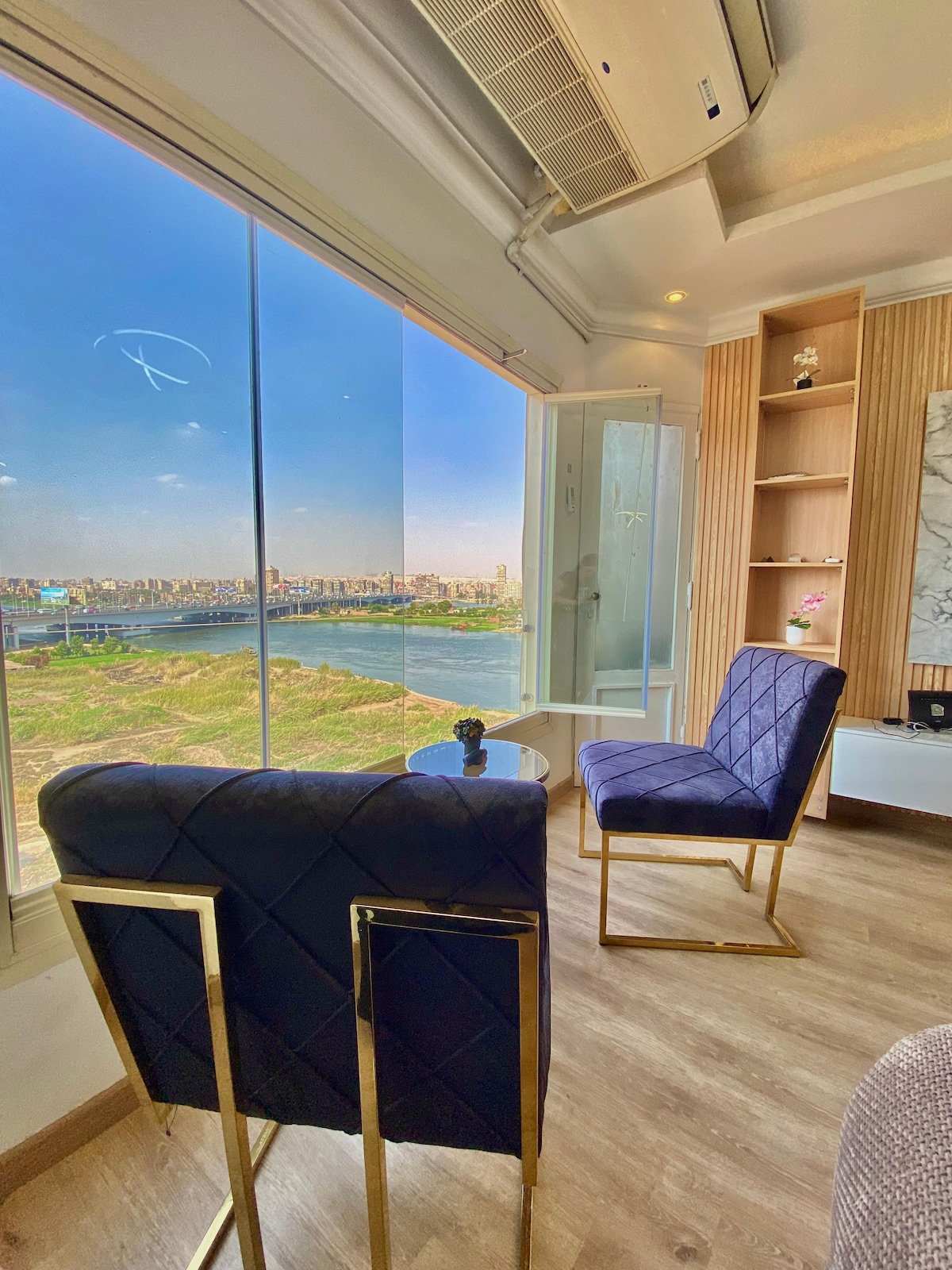 A cozy seating area is presented with two elegant chairs in deep blue upholstery and gold accents. A small table sits between them, accompanied by a decorative plant. Large windows offer expansive views of the Nile and the surrounding landscape.