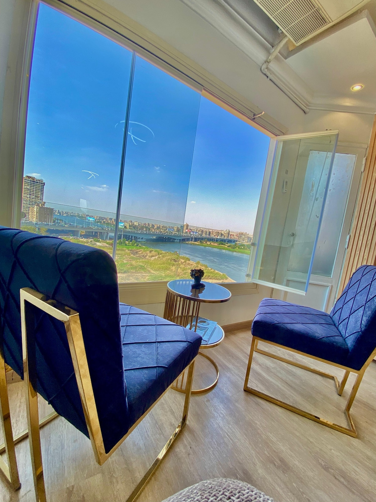 Two elegant chairs with gold frames are positioned near a window, providing a view of the river. A small round table is situated between them, while natural light enters the space, enhancing the serene atmosphere of the corner seating area.