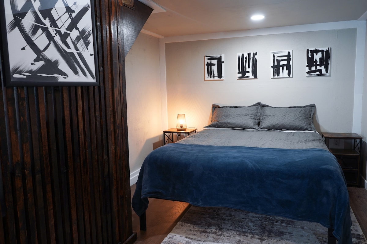 A serene bedroom with a large queen-size bed, modern wall art, warm wood accents, and a bedside table, creating a calm, stylish, and inviting retreat.
