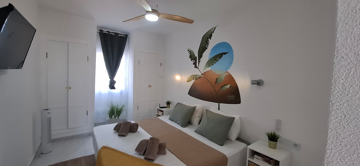 A bright bedroom features a spacious bed dressed with neutral linens and decorative towels. A wall-mounted television is positioned across from the bed. Natural light filters in through a window framed by dark curtains, while a wall mural of leaves adds a touch of nature.