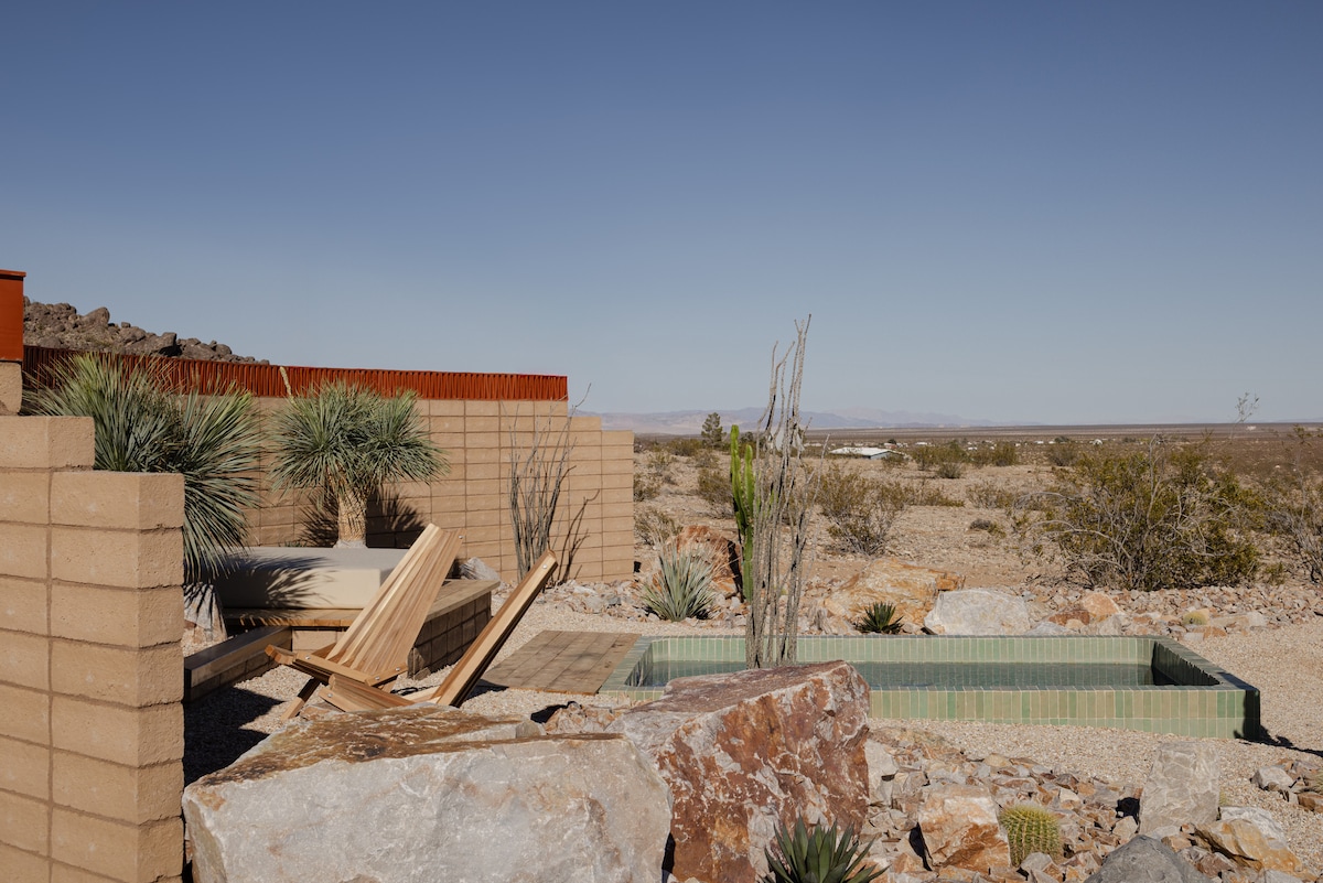 An outdoor space features a private plunge pool, woven sun bed, and natural stone accents. Desert plants and rock formations enhance the tranquil setting, with open desert views stretching into the distance beneath a clear blue sky.