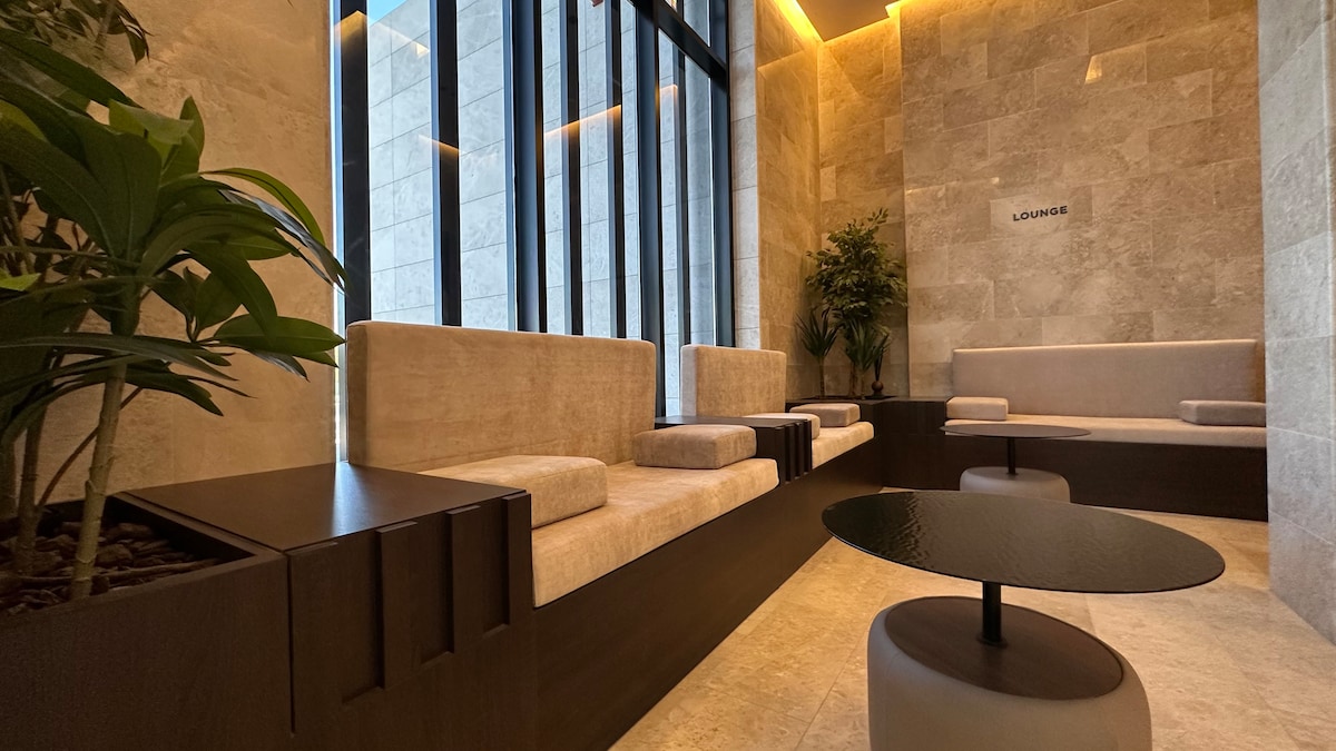 A modern lounge area is presented with soft, beige seating arranged around a sleek, dark coffee table. Large windows allow natural light to illuminate the space, complemented by plants that add a touch of greenery to the neutral color palette.