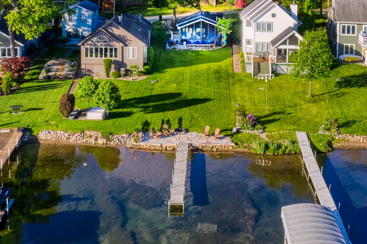 A sunny lakeside view captures well-maintained lawns and multiple cottages along the water. A wooden dock extends into the lake, providing access for water activities. Lush greenery is complemented by colorful flowers, enhancing the serene atmosphere of this family-friendly environment.
