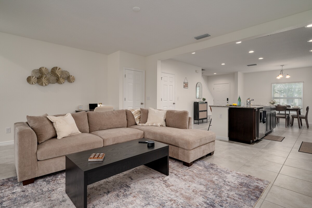 A spacious living area features a comfortable sectional sofa and a rectangular coffee table, set on a soft area rug. The open layout connects to a dining space and a modern kitchen, providing a welcoming environment for gatherings.