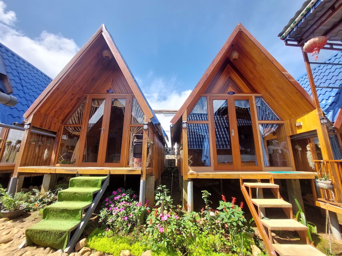 Two wooden bungalows are positioned side by side, each featuring large glass doors that open onto a small outdoor area. The roofs are sharply angled, and a landscaped path with vibrant flowers runs in front of the structures, enhancing the inviting atmosphere.