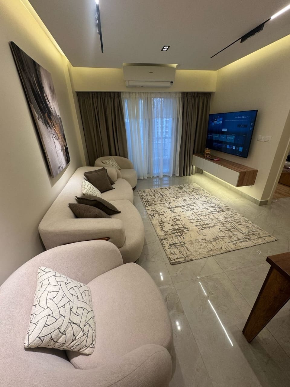 A softly lit living area features a light-colored sectional sofa adorned with decorative cushions. A wall-mounted TV is positioned above a sleek, minimalistic console. A large window covered with sheer curtains allows natural light to fill the space, while a patterned rug adds warmth.