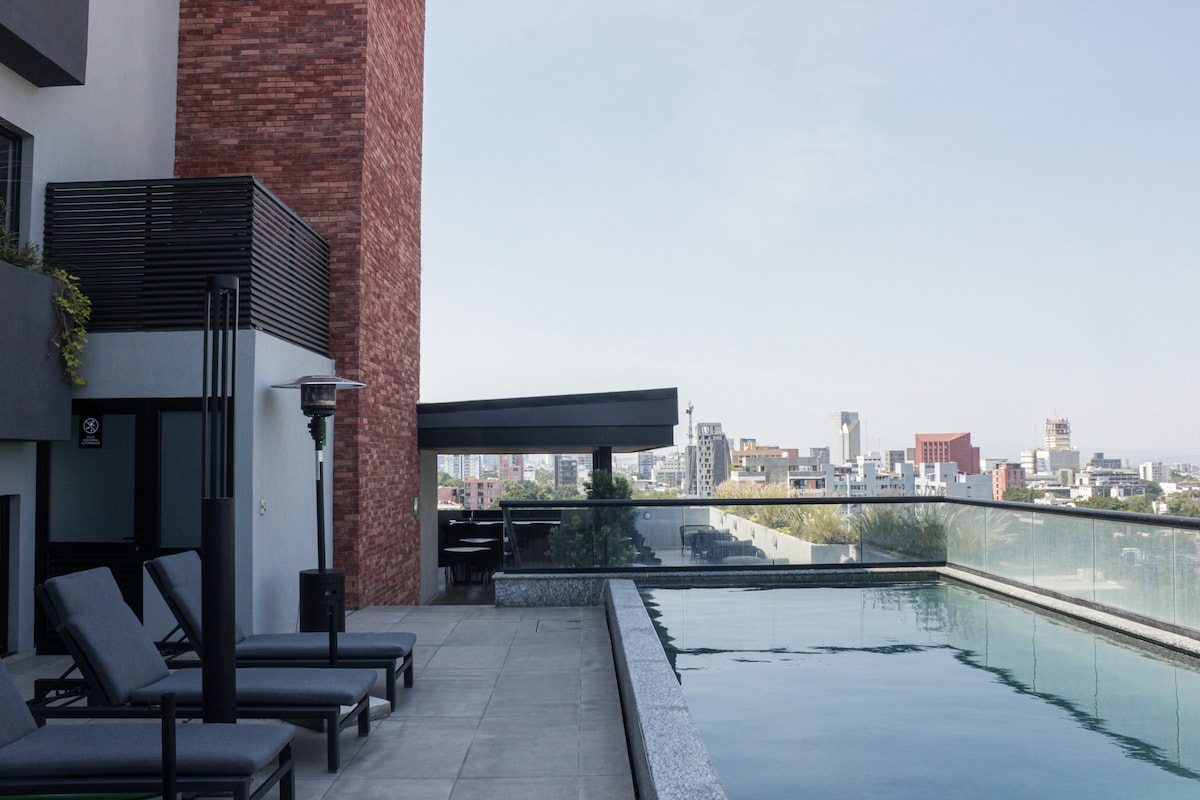 An outdoor pool area features sleek loungers arranged alongside a modern infinity pool overlooking the city skyline. Clear glass railings provide unobstructed views, while comfortable seating is positioned under a shaded structure, promoting relaxation in an inviting environment.