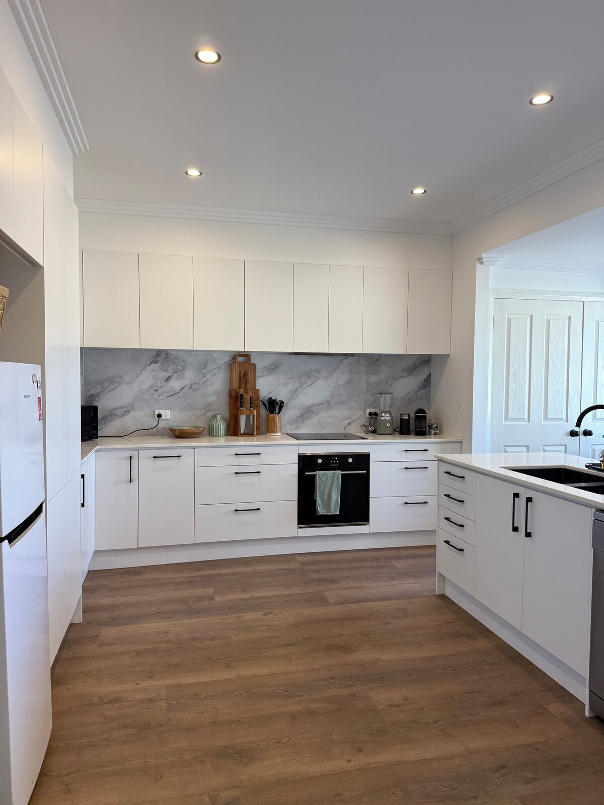 A modern kitchen is displayed, featuring white cabinetry and a sleek marble backsplash. Stainless steel appliances, including an oven and refrigerator, complement the clean design. Natural light fills the space through an adjacent entrance, enhancing the overall bright and functional layout.