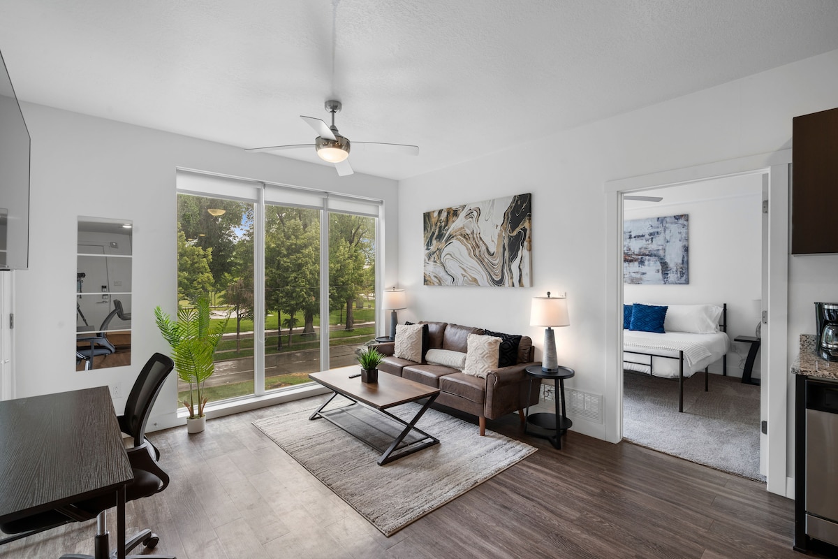 A modern living area showcases a comfortable sofa paired with a stylish coffee table. Large windows allow natural light to brighten the space, while greenery from outside complements the neutral color palette. A workspace with a chair and a second bedroom is visible through the doorway.